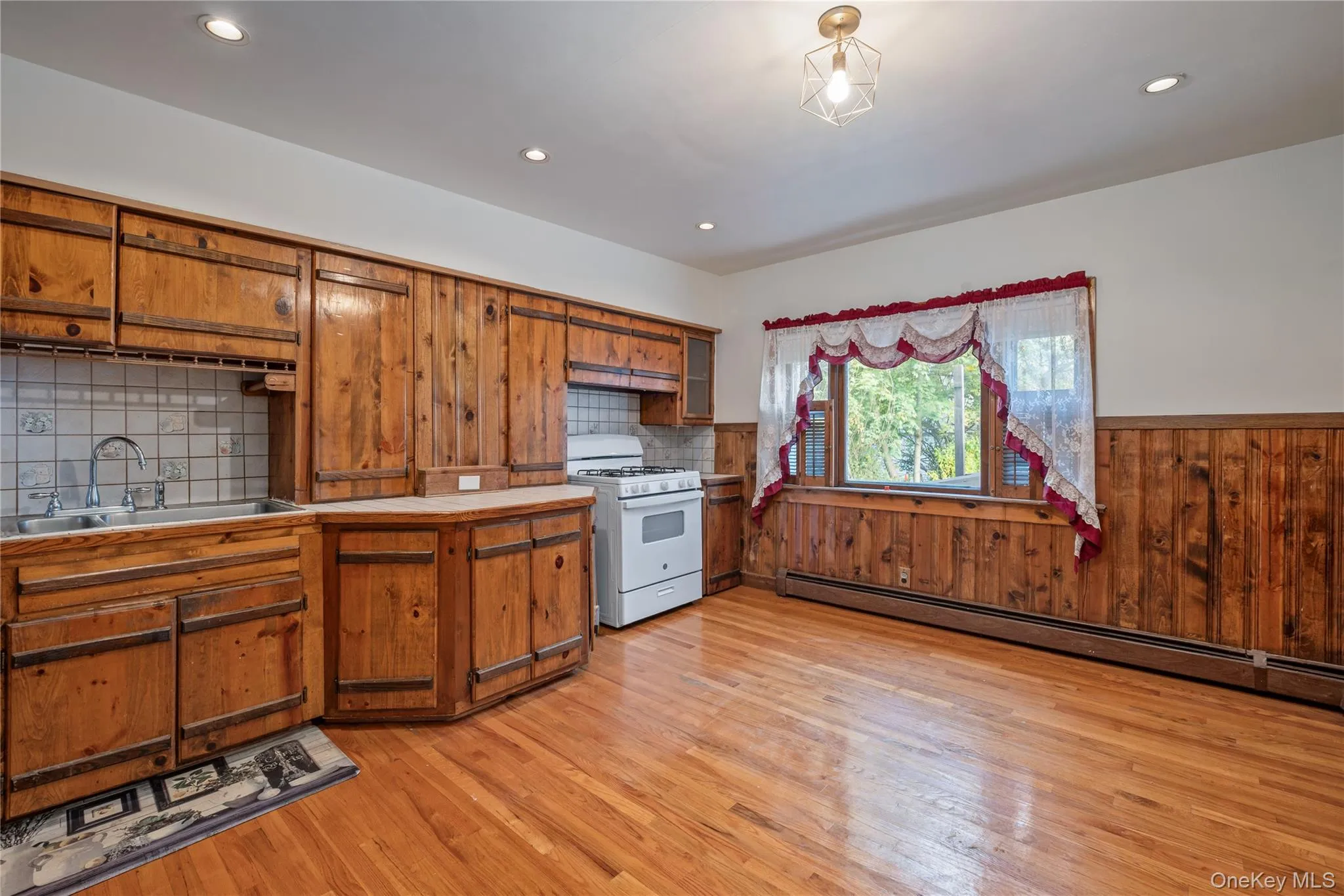 Kitchen with brown cabinetry, white range with gas stovetop, light wood-style floors, a baseboard heating unit, and wood walls Kitchen with brown cabinetry, white range with gas stovetop, light wood-style floors, a baseboard heating unit, and wood walls