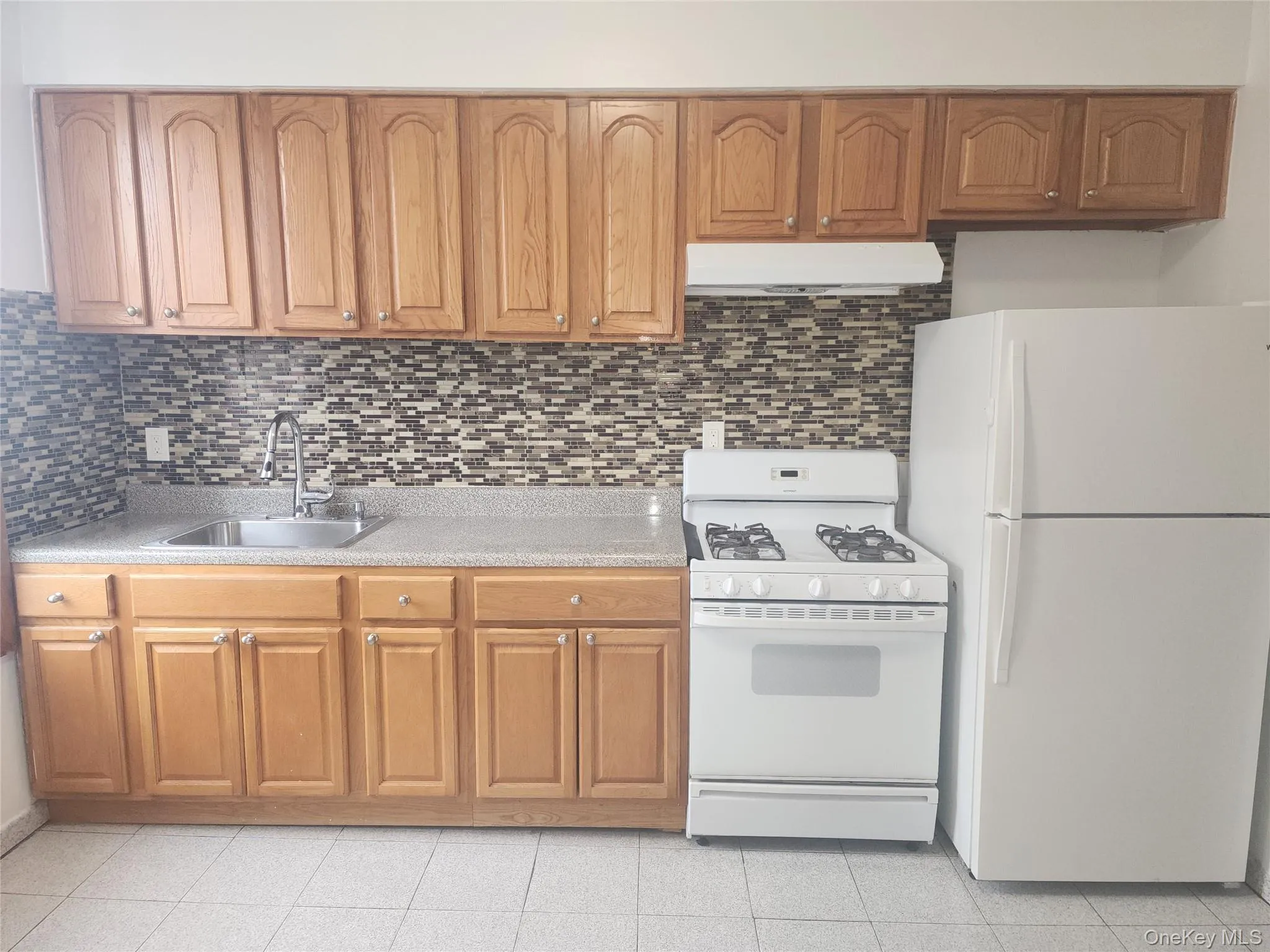 Kitchen featuring white appliances, brown cabinetry, tasteful backsplash, and under cabinet range hood Kitchen featuring white appliances, brown cabinetry, tasteful backsplash, and under cabinet range hood