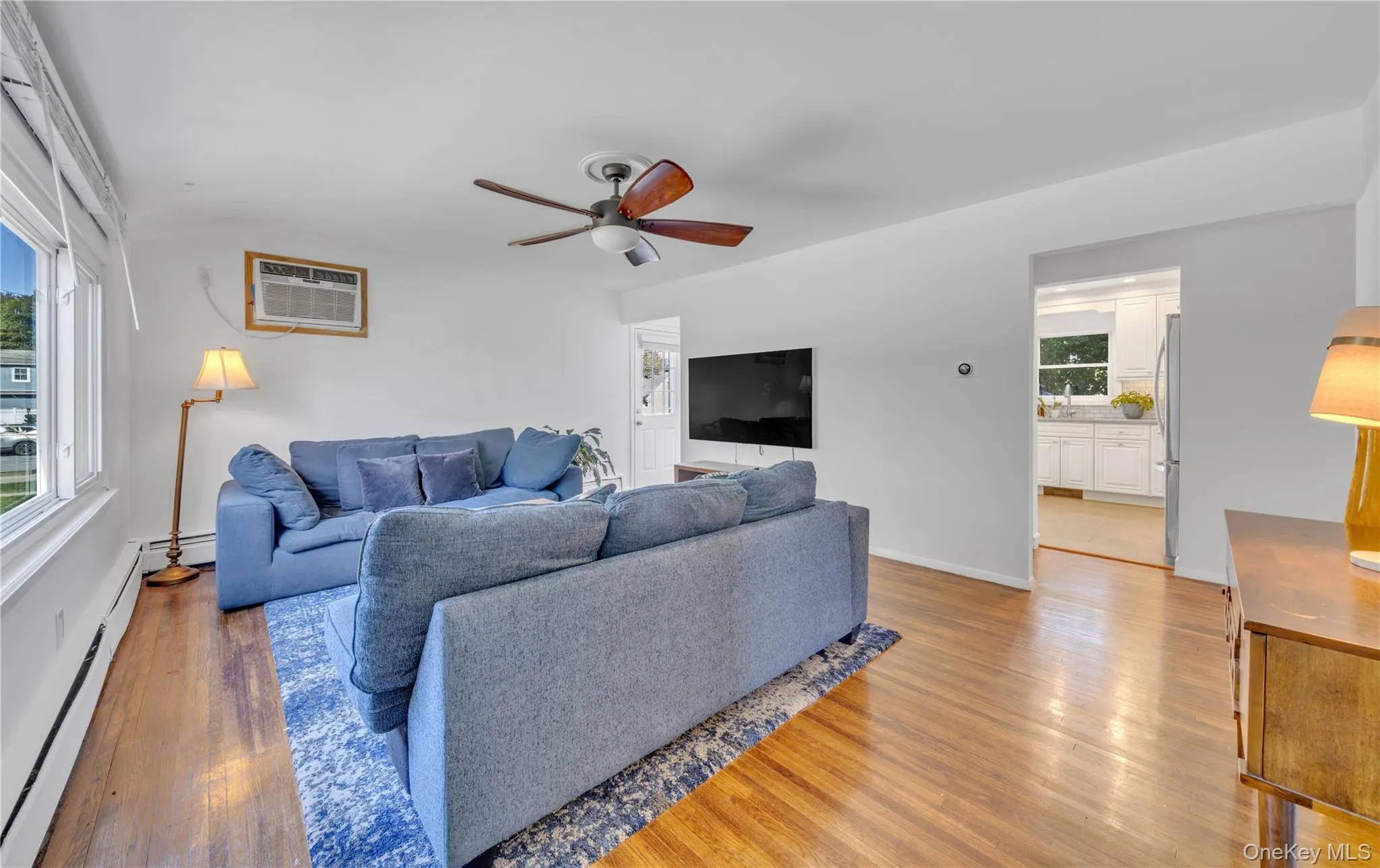 Living room with light wood-style flooring, a ceiling fan, an AC wall unit, and a baseboard heating unit Living room with light wood-style flooring, a ceiling fan, an AC wall unit, and a baseboard heating unit