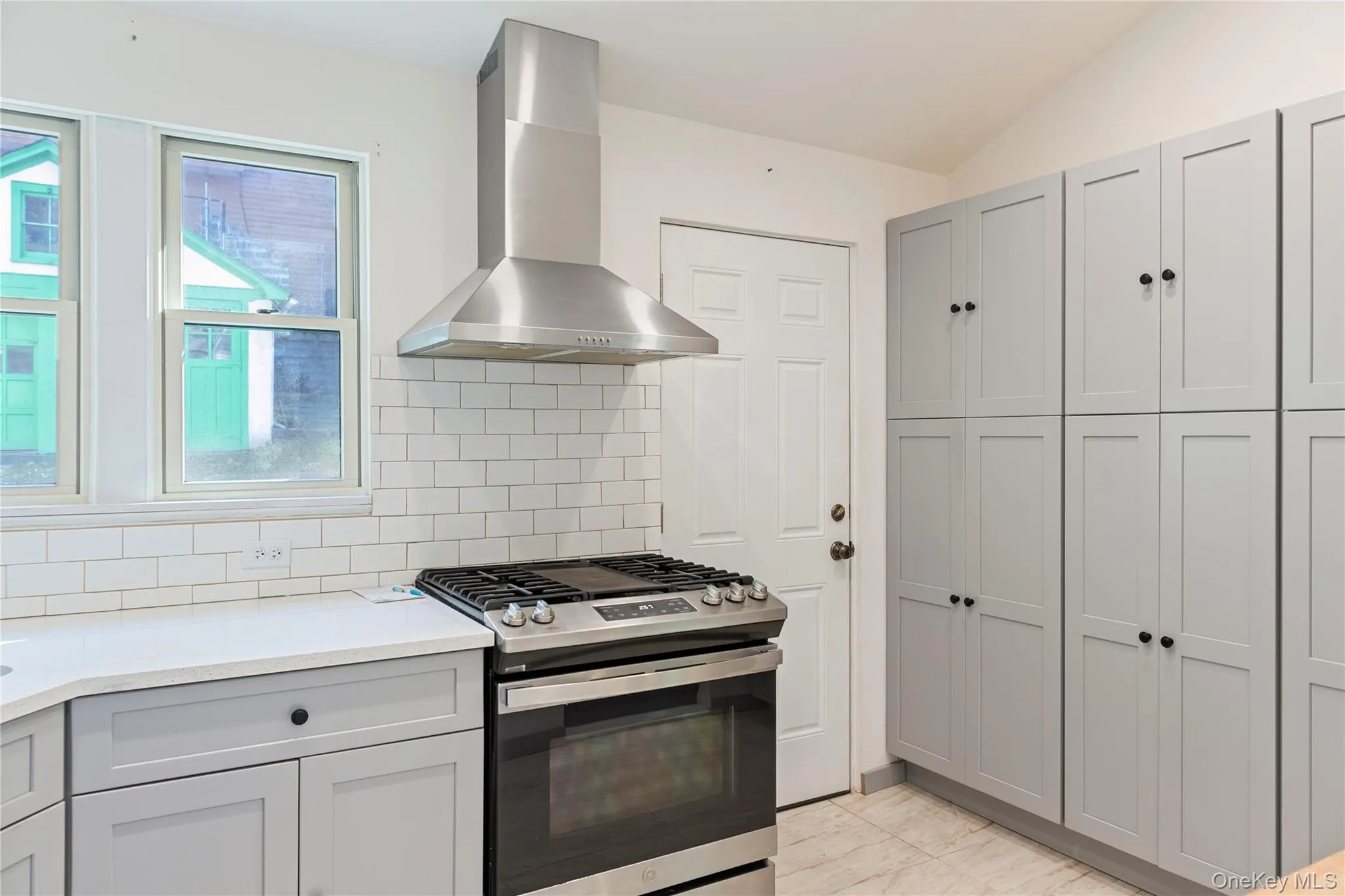 Kitchen with gray cabinets, stainless steel gas stove, wall chimney range hood, decorative backsplash, and light stone counters Kitchen with gray cabinets, stainless steel gas stove, wall chimney range hood, decorative backsplash, and light stone counters