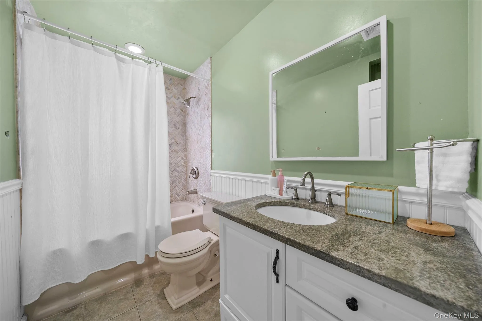 Full bathroom featuring shower / bathtub combination with curtain, vanity, light tile patterned floors, and a wainscoted wall Full bathroom featuring shower / bathtub combination with curtain, vanity, light tile patterned floors, and a wainscoted wall