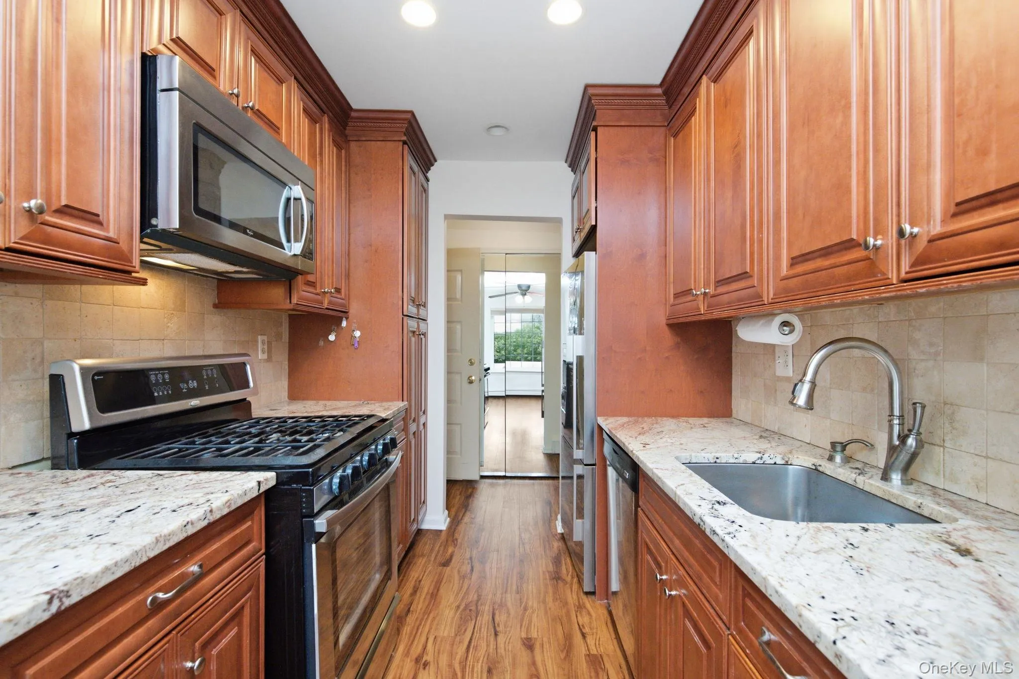 The galley kitchen features rich wood cabinetry with crown molding detail, granite countertops, a tile backsplash, and stainless steel appliances including a gas range and built-in microwave. The galley kitchen features rich wood cabinetry with crown molding detail, granite countertops, a tile backsplash, and stainless steel appliances including a gas range and built-in microwave.