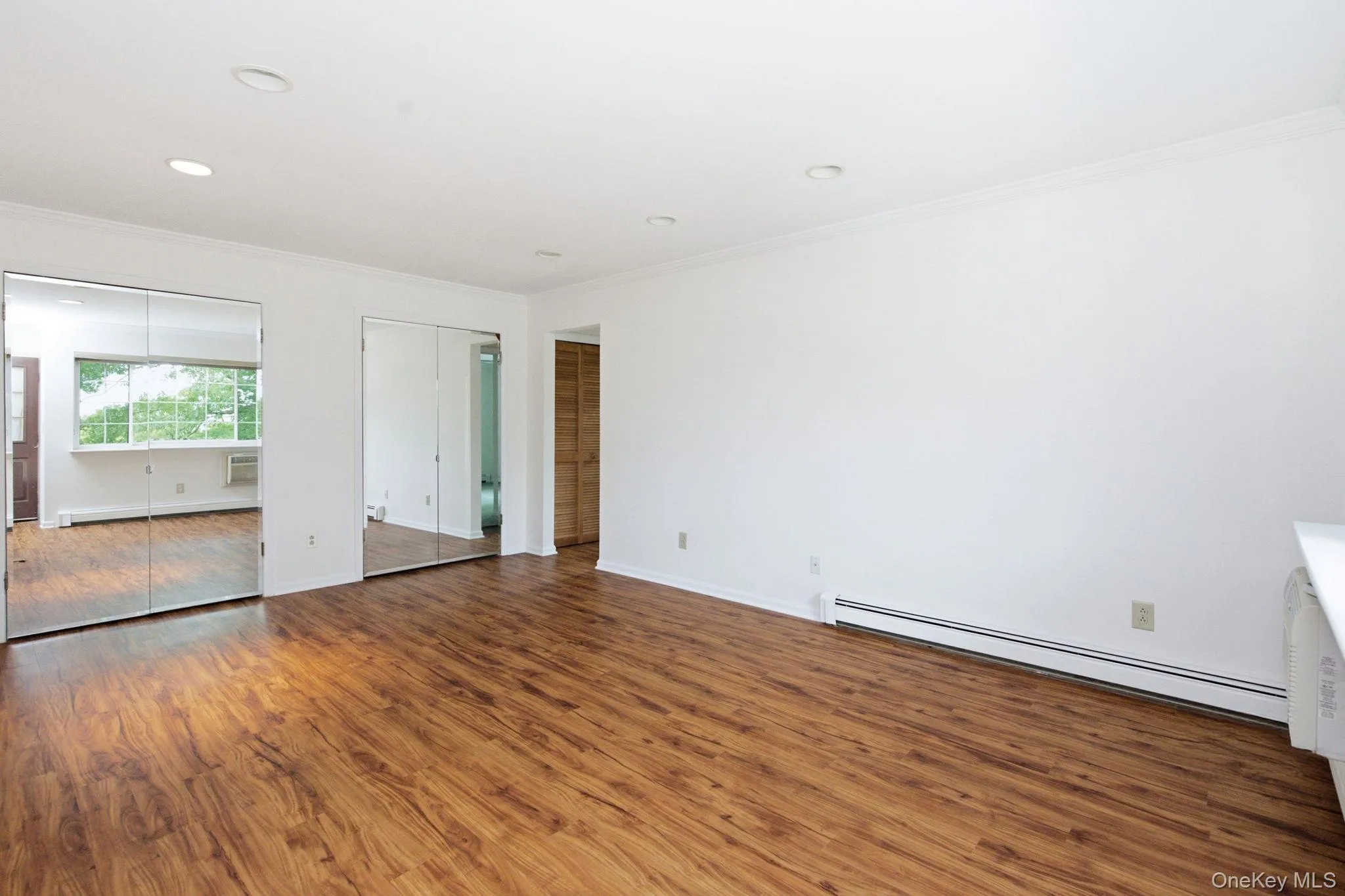 The living space flows toward the bedroom through an open corridor, with mirror-paneled closet doors reflecting light, recessed fixtures above, and louvered laundry doors just off to the side. The living space flows toward the bedroom through an open corridor, with mirror-paneled closet doors reflecting light, recessed fixtures above, and louvered laundry doors just off to the side.