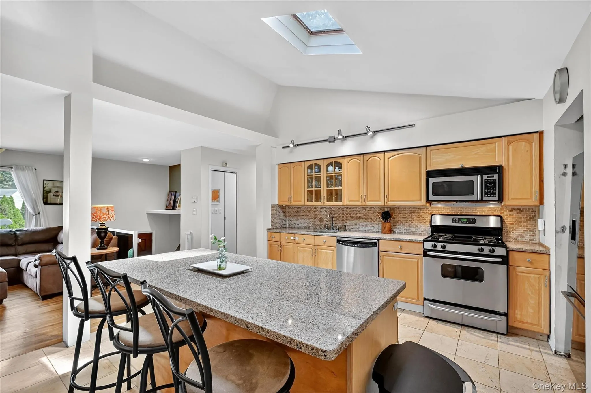 Kitchen featuring a skylight, appliances with stainless steel finishes, a kitchen bar, glass insert cabinets, and backsplash Kitchen featuring a skylight, appliances with stainless steel finishes, a kitchen bar, glass insert cabinets, and backsplash