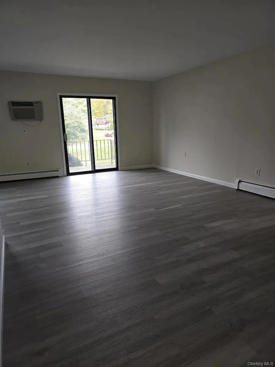 Empty room featuring dark wood-style flooring, baseboard heating, and an AC wall unit Empty room featuring dark wood-style flooring, baseboard heating, and an AC wall unit