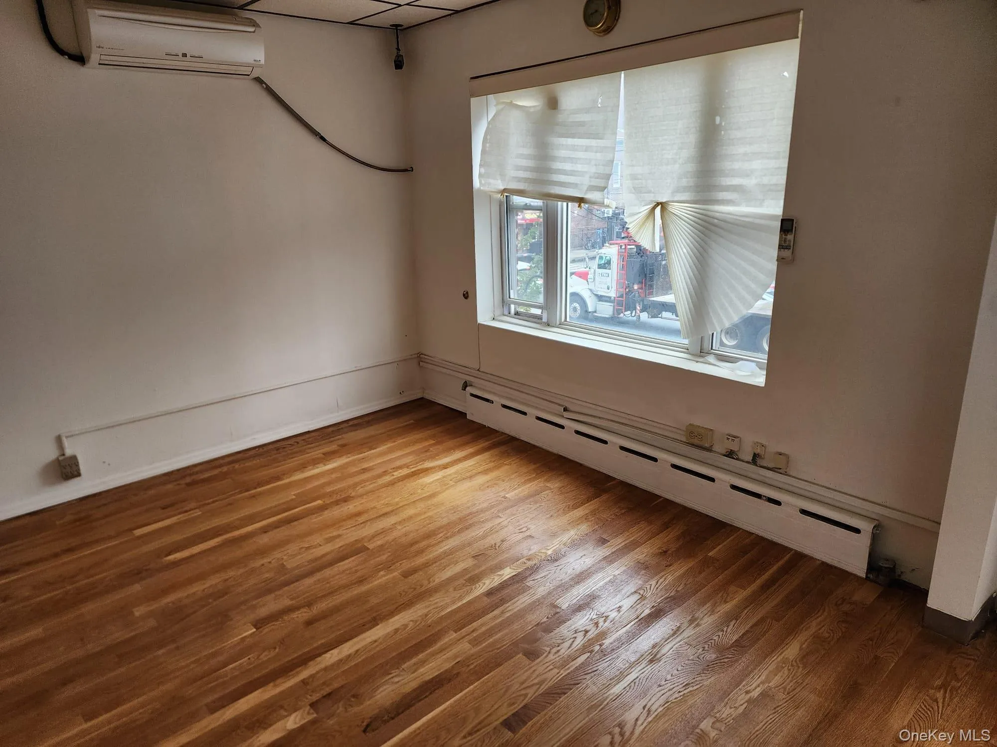 Unfurnished room with a baseboard heating unit, a wall mounted AC, wood finished floors, and a drop ceiling Unfurnished room with a baseboard heating unit, a wall mounted AC, wood finished floors, and a drop ceiling