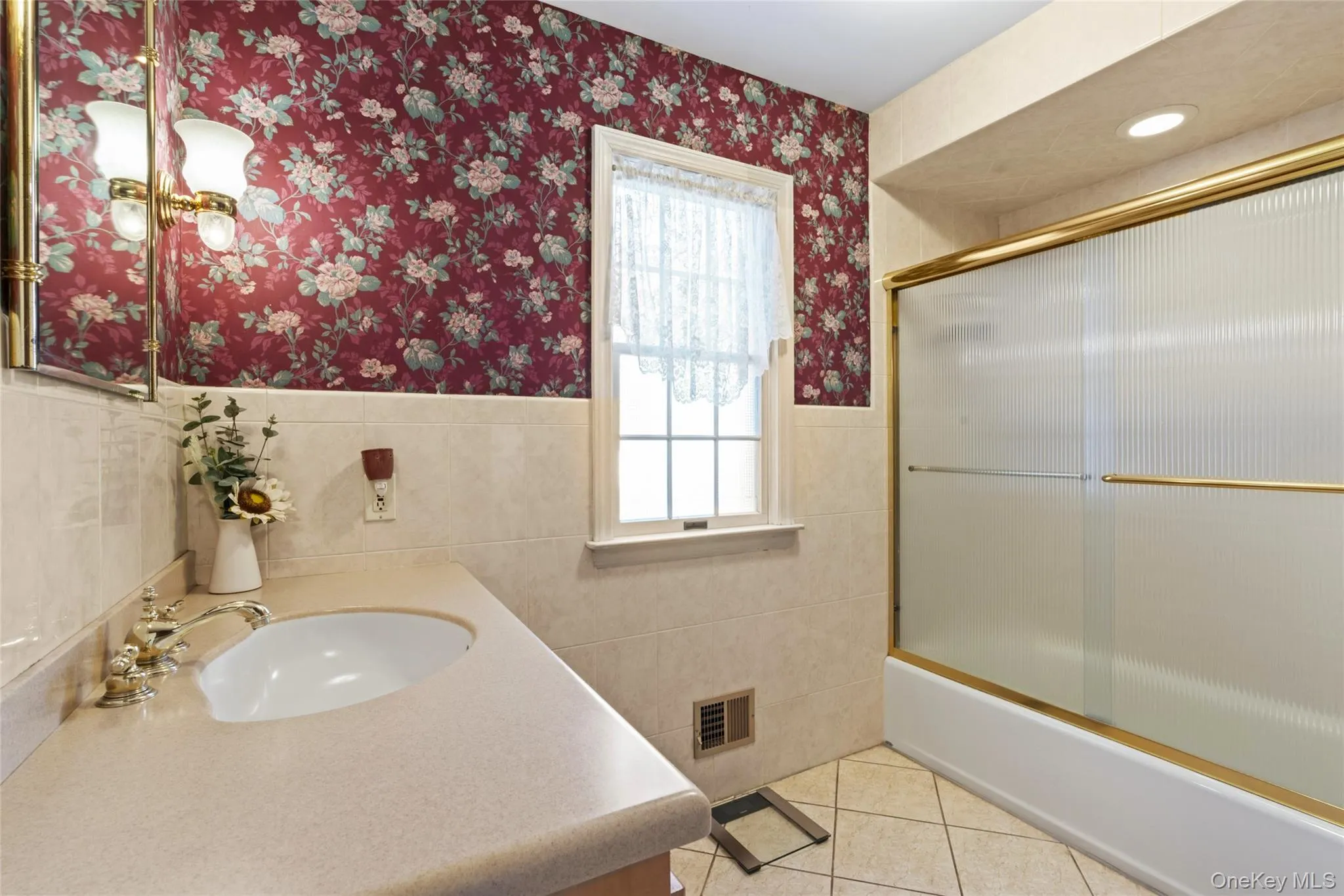 Bathroom featuring vanity, tile walls, shower / bath combination with glass door, light tile patterned flooring, and wallpapered walls Bathroom featuring vanity, tile walls, shower / bath combination with glass door, light tile patterned flooring, and wallpapered walls
