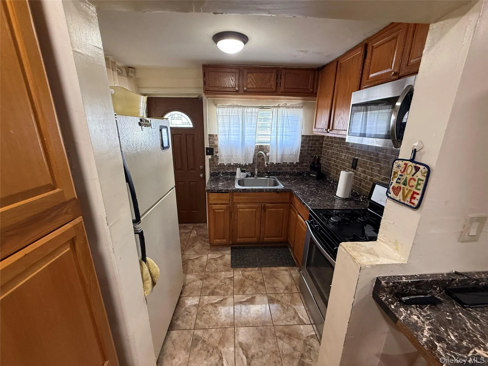 Kitchen featuring gas stove, stainless steel microwave, brown cabinetry, backsplash, and freestanding refrigerator Kitchen featuring gas stove, stainless steel microwave, brown cabinetry, backsplash, and freestanding refrigerator