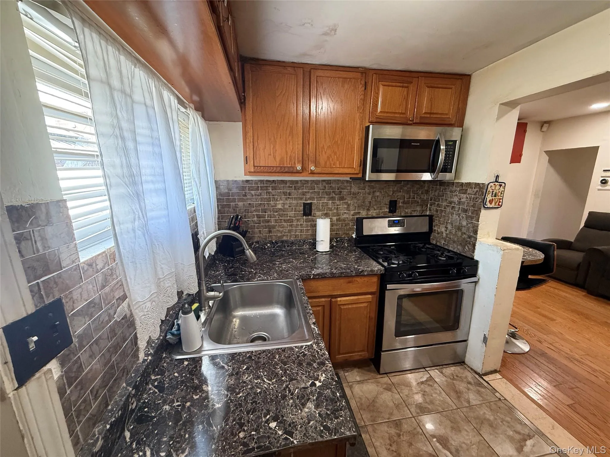 Kitchen with brown cabinets, tasteful backsplash, stainless steel appliances, dark countertops, and light tile patterned floors Kitchen with brown cabinets, tasteful backsplash, stainless steel appliances, dark countertops, and light tile patterned floors