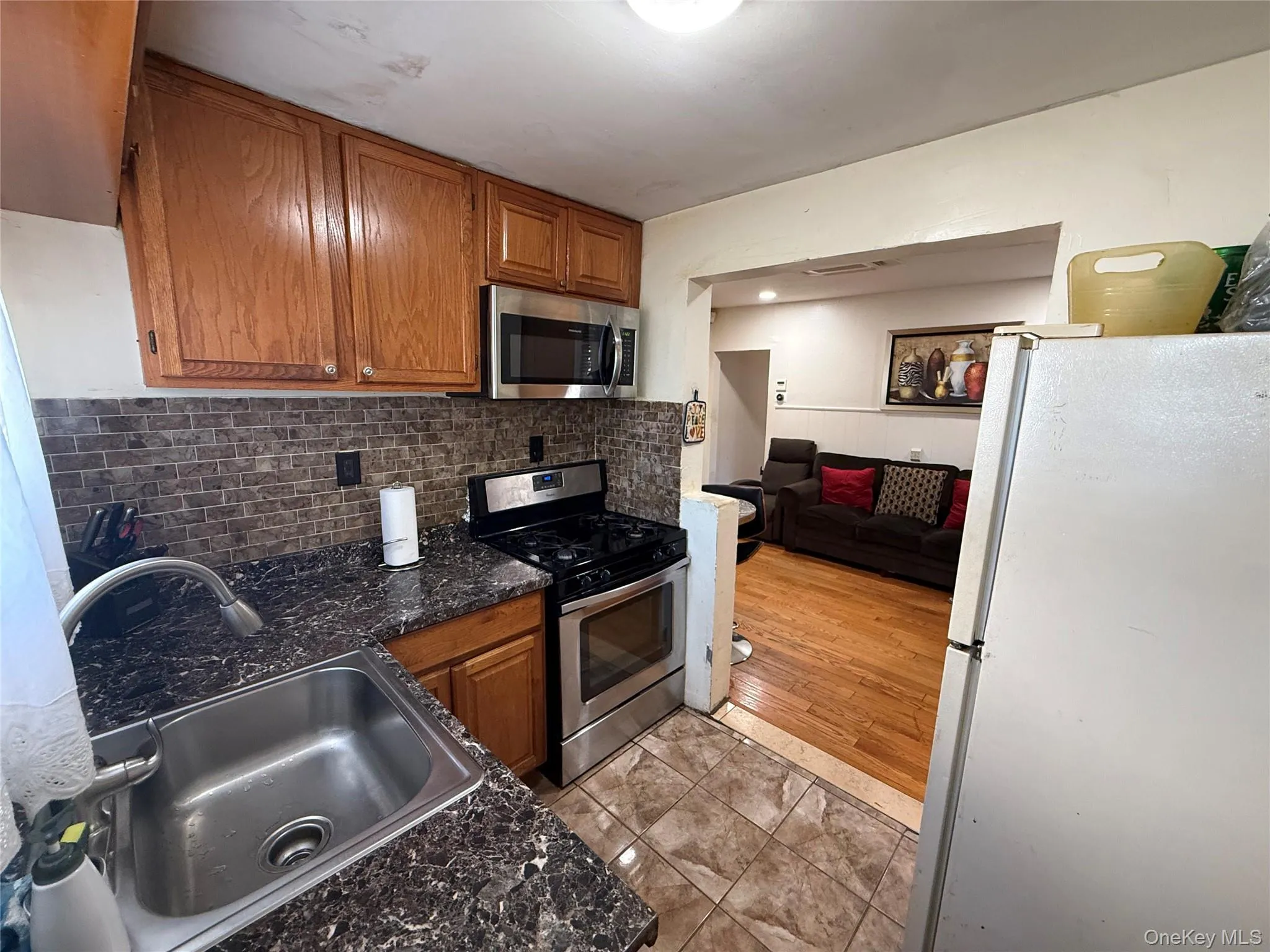 Kitchen featuring stainless steel appliances, brown cabinetry, decorative backsplash, light tile patterned flooring, and dark stone counters Kitchen featuring stainless steel appliances, brown cabinetry, decorative backsplash, light tile patterned flooring, and dark stone counters