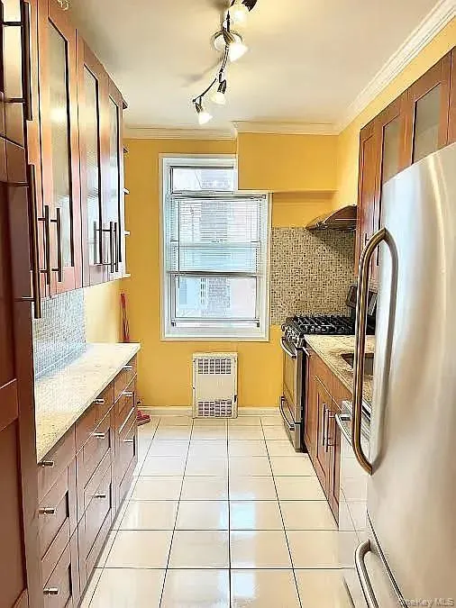Kitchen featuring tasteful backsplash, brown cabinets, stainless steel appliances, light tile patterned flooring, and crown molding Kitchen featuring tasteful backsplash, brown cabinets, stainless steel appliances, light tile patterned flooring, and crown molding