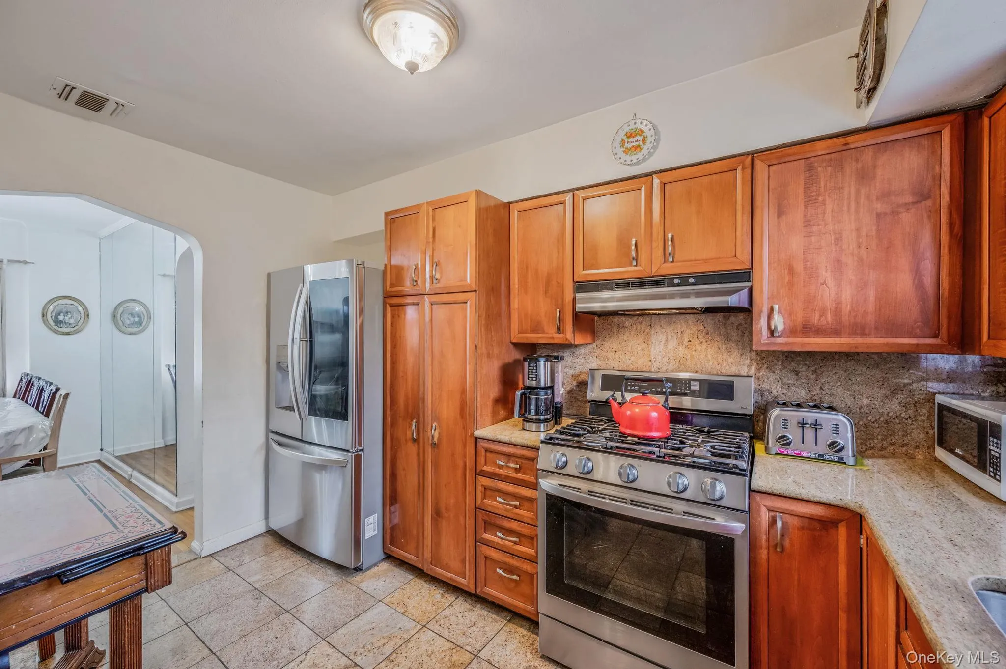 Kitchen featuring stainless steel appliances, brown cabinetry, backsplash, arched walkways, and under cabinet range hood Kitchen featuring stainless steel appliances, brown cabinetry, backsplash, arched walkways, and under cabinet range hood