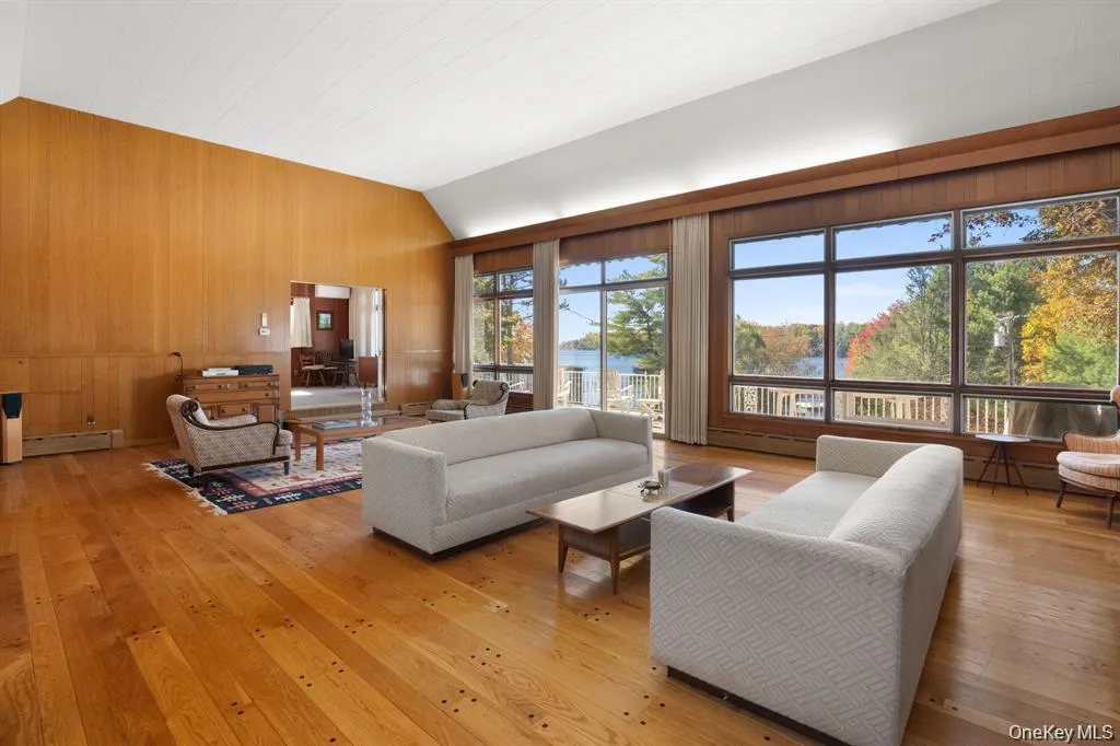 Living room featuring wooden walls, light wood-style flooring, vaulted ceiling, a baseboard heating unit, and a water view Living room featuring wooden walls, light wood-style flooring, vaulted ceiling, a baseboard heating unit, and a water view
