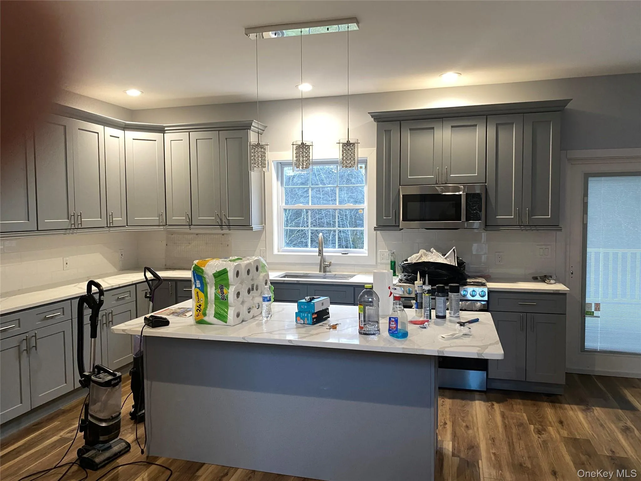 Kitchen featuring gray cabinetry, a center island with sink, stainless steel microwave, hanging light fixtures, and light stone counters Kitchen featuring gray cabinetry, a center island with sink, stainless steel microwave, hanging light fixtures, and light stone counters