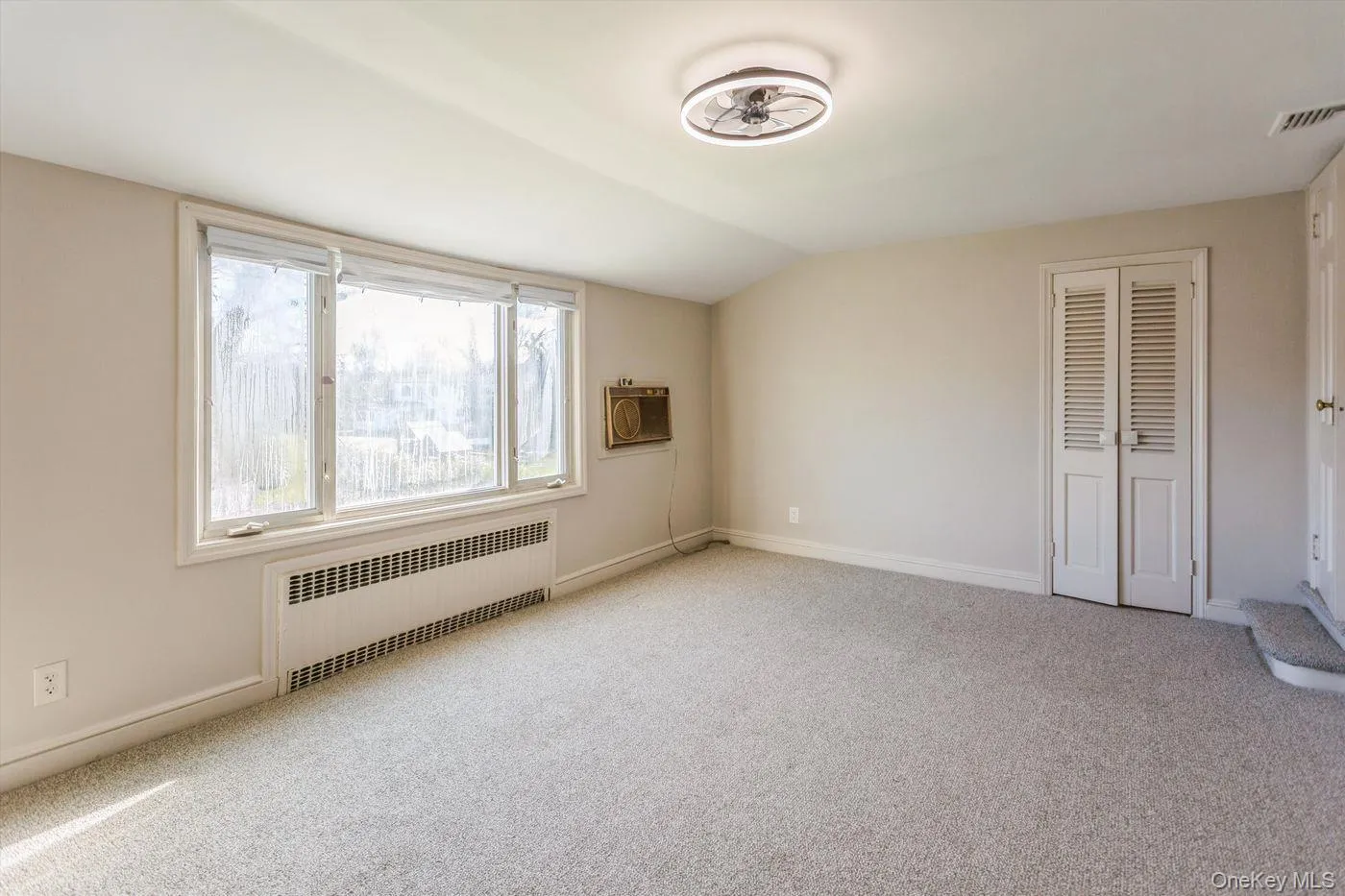 Empty room featuring radiator, light colored carpet, lofted ceiling, and an AC wall unit Empty room featuring radiator, light colored carpet, lofted ceiling, and an AC wall unit