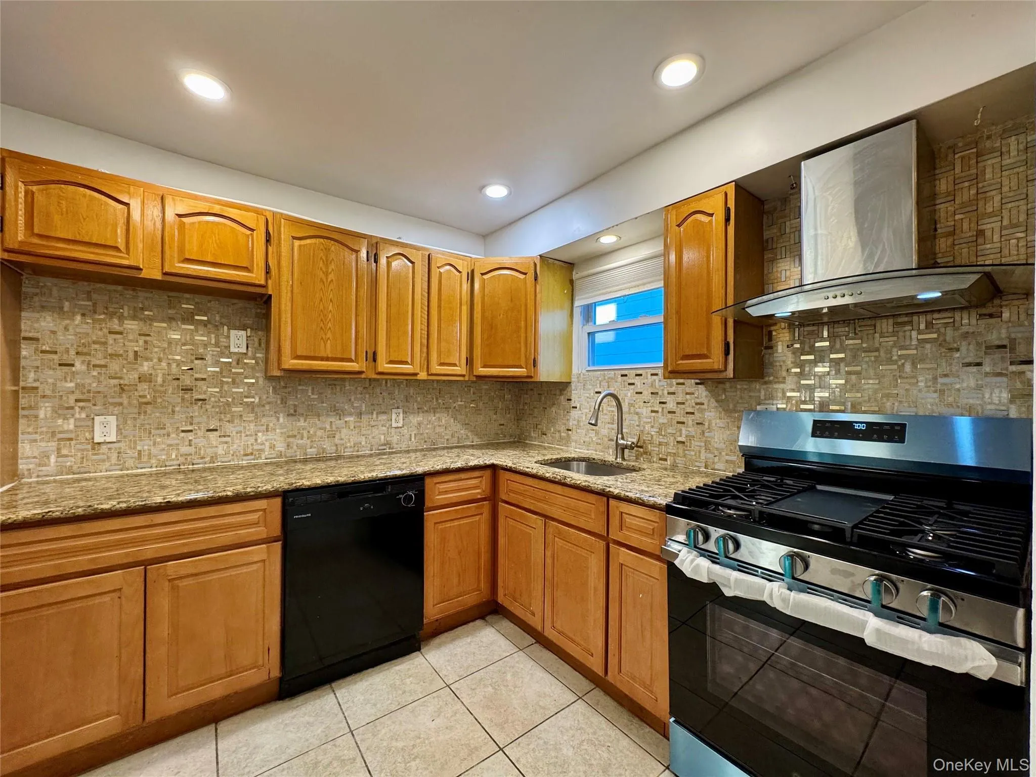 Kitchen featuring stainless steel gas range oven, tasteful backsplash, light stone countertops, wall chimney range hood, and brown cabinets Kitchen featuring stainless steel gas range oven, tasteful backsplash, light stone countertops, wall chimney range hood, and brown cabinets