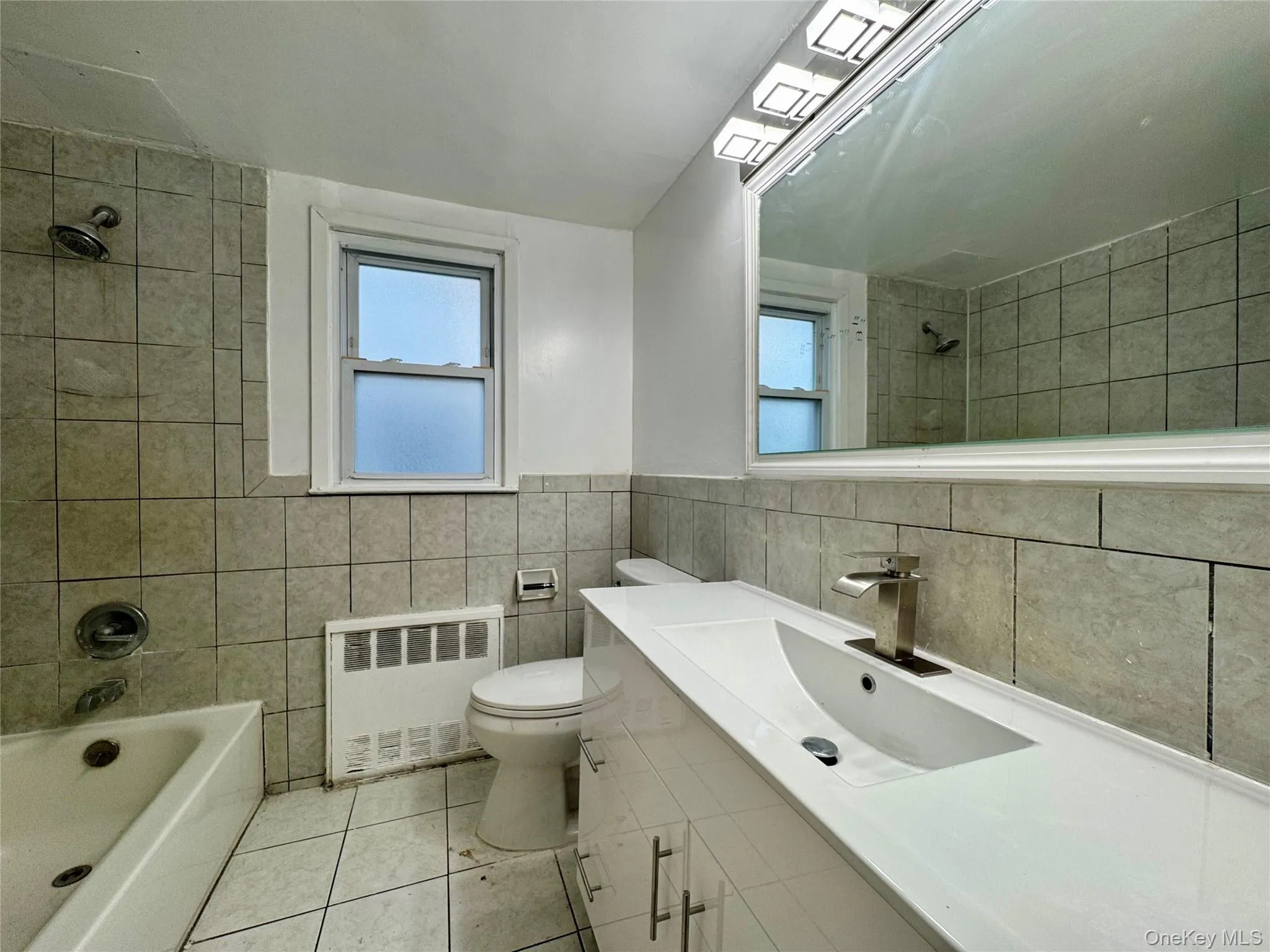Full bath featuring radiator, tile walls, vanity, light tile patterned floors, and healthy amount of natural light Full bath featuring radiator, tile walls, vanity, light tile patterned floors, and healthy amount of natural light