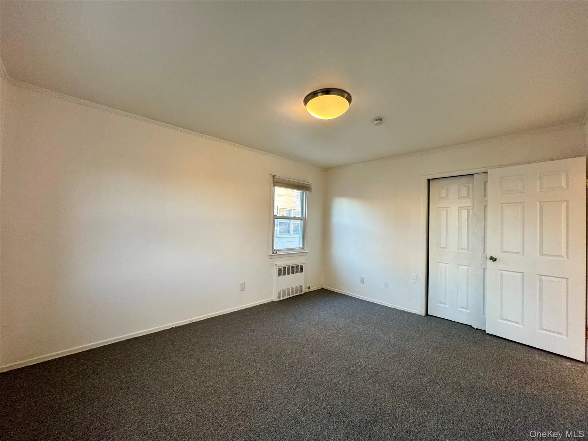 Unfurnished bedroom with dark colored carpet, radiator heating unit, a closet, and crown molding Unfurnished bedroom with dark colored carpet, radiator heating unit, a closet, and crown molding