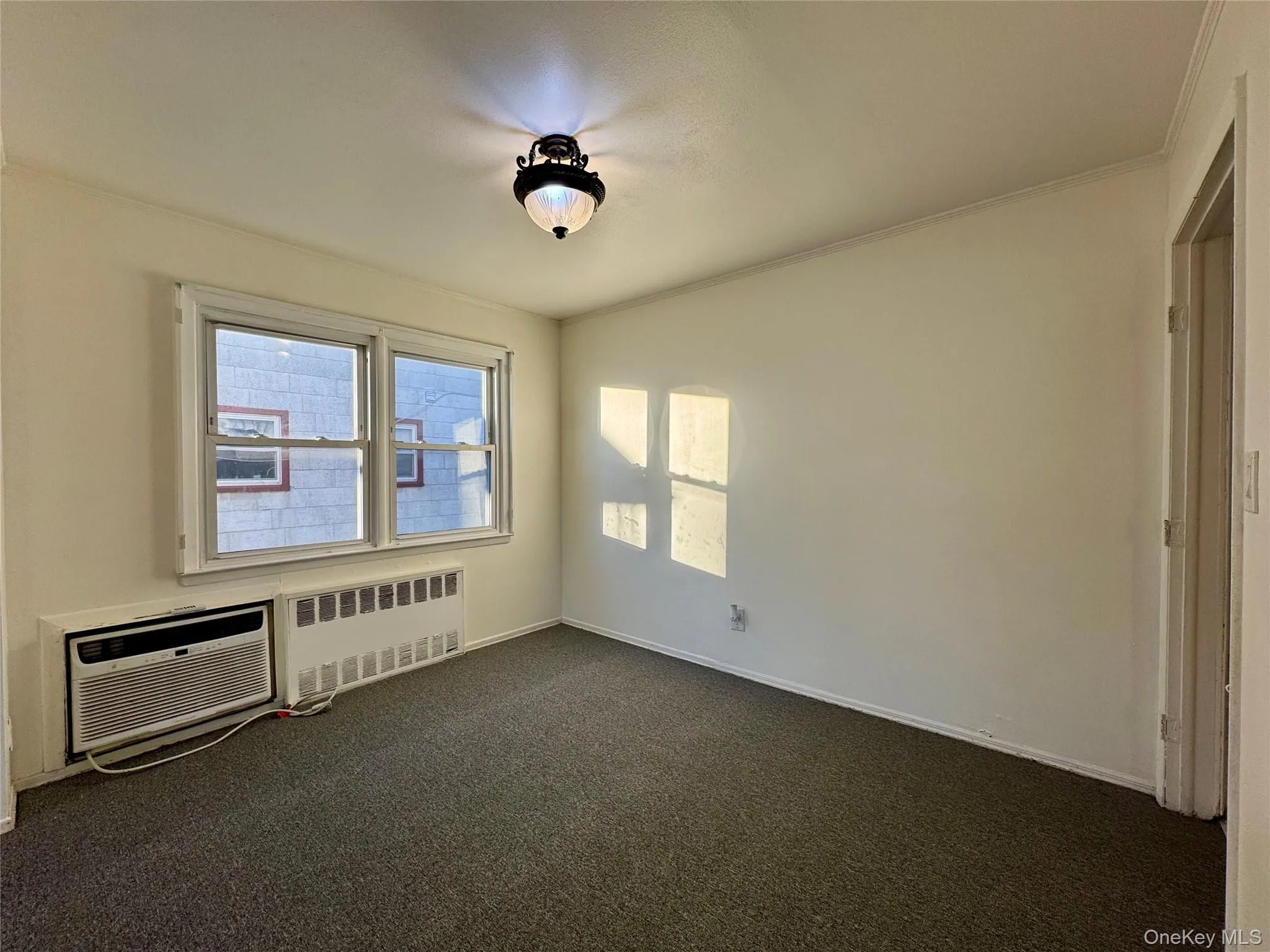 Empty room with radiator, plenty of natural light, dark colored carpet, an AC wall unit, and ornamental molding Empty room with radiator, plenty of natural light, dark colored carpet, an AC wall unit, and ornamental molding