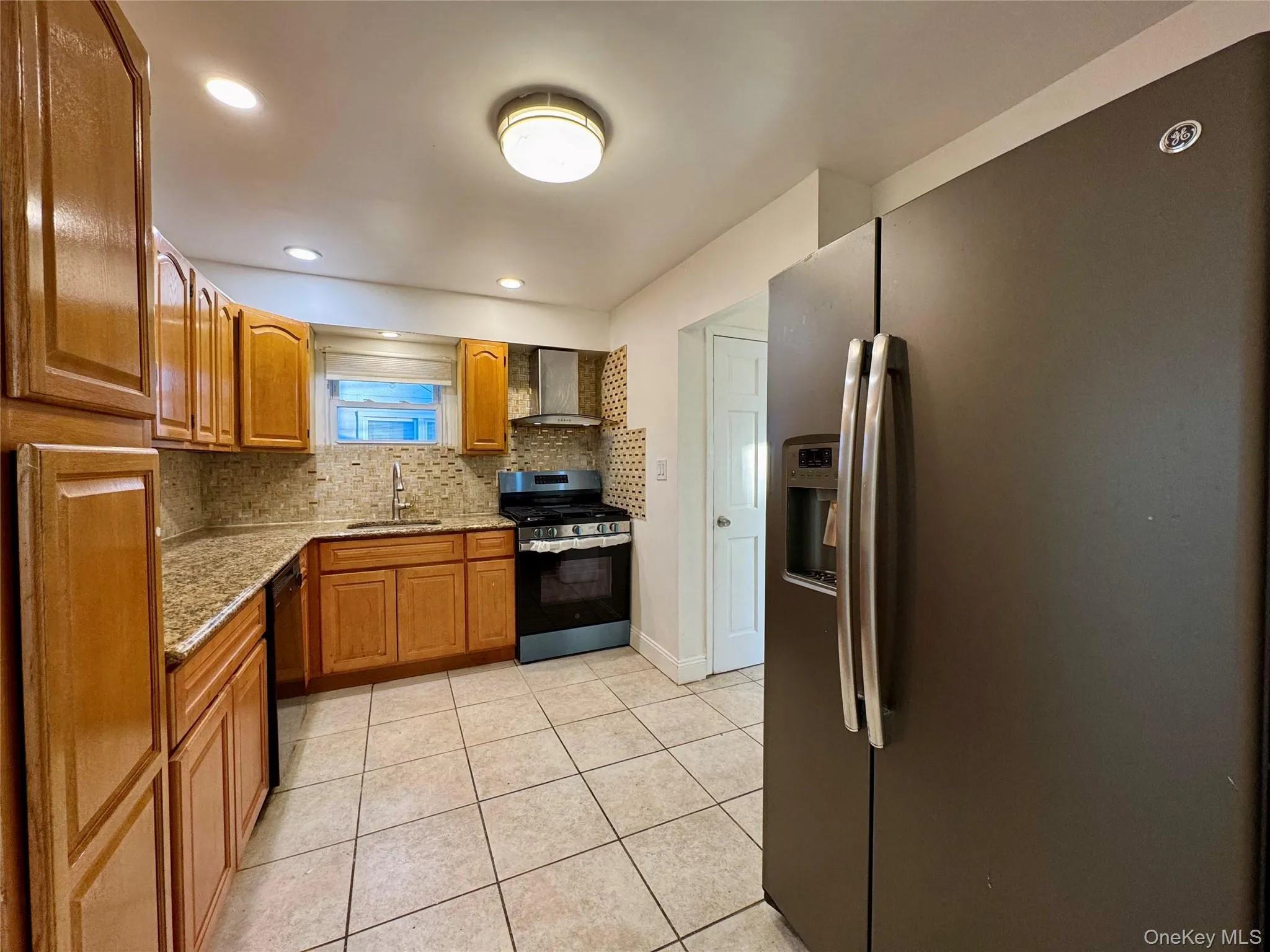 Kitchen featuring appliances with stainless steel finishes, light stone counters, brown cabinetry, decorative backsplash, and wall chimney range hood Kitchen featuring appliances with stainless steel finishes, light stone counters, brown cabinetry, decorative backsplash, and wall chimney range hood