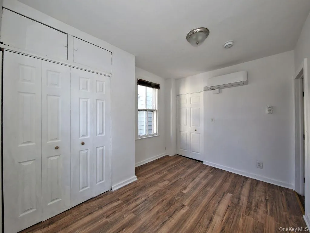Unfurnished bedroom with two closets, dark wood-style flooring, and an AC wall unit Unfurnished bedroom with two closets, dark wood-style flooring, and an AC wall unit