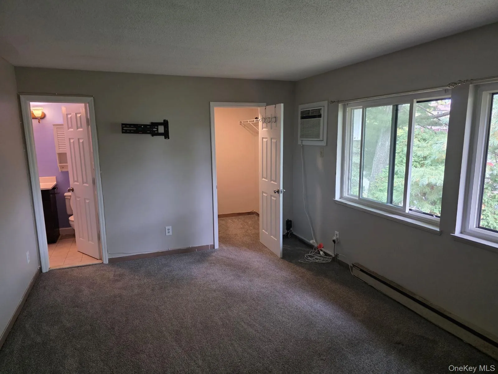 Unfurnished bedroom with a baseboard radiator, a spacious closet, carpet floors, a textured ceiling, and an AC wall unit Unfurnished bedroom with a baseboard radiator, a spacious closet, carpet floors, a textured ceiling, and an AC wall unit