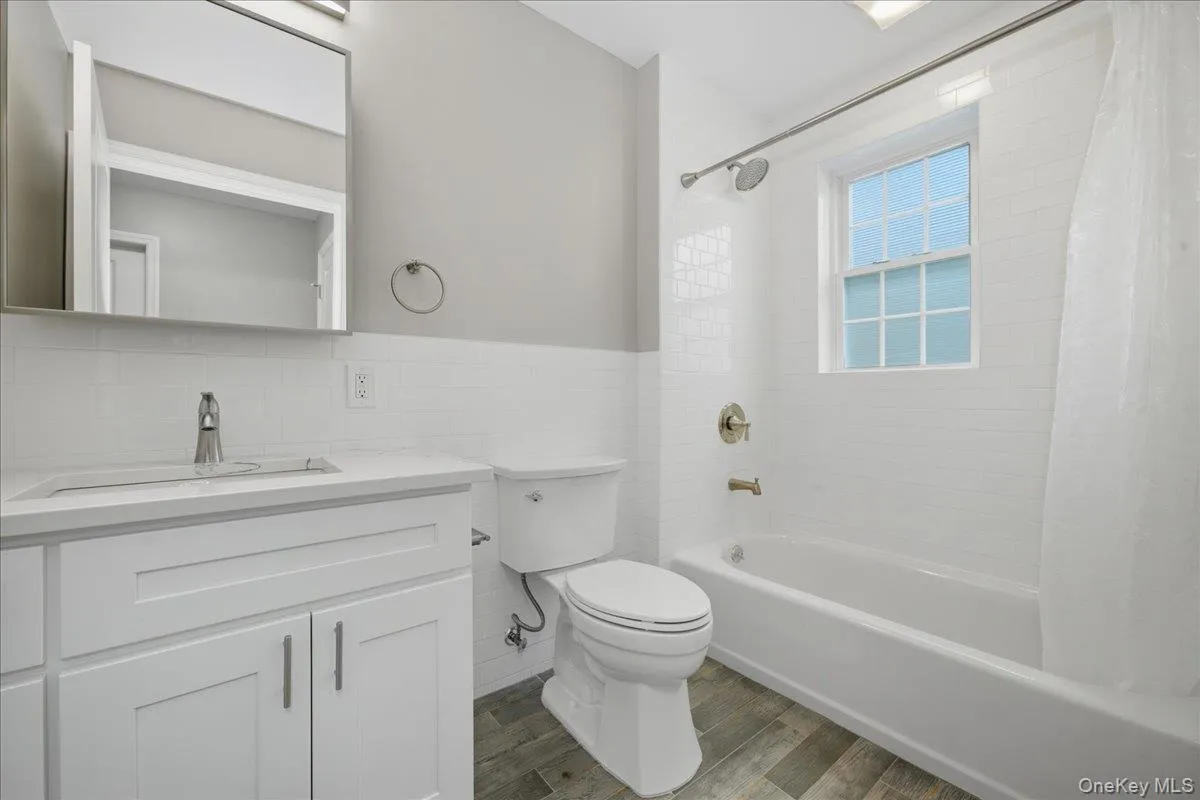 Full bath with shower / bathtub combination with curtain, vanity, dark wood-style flooring, tile walls, and a wainscoted wall Full bath with shower / bathtub combination with curtain, vanity, dark wood-style flooring, tile walls, and a wainscoted wall