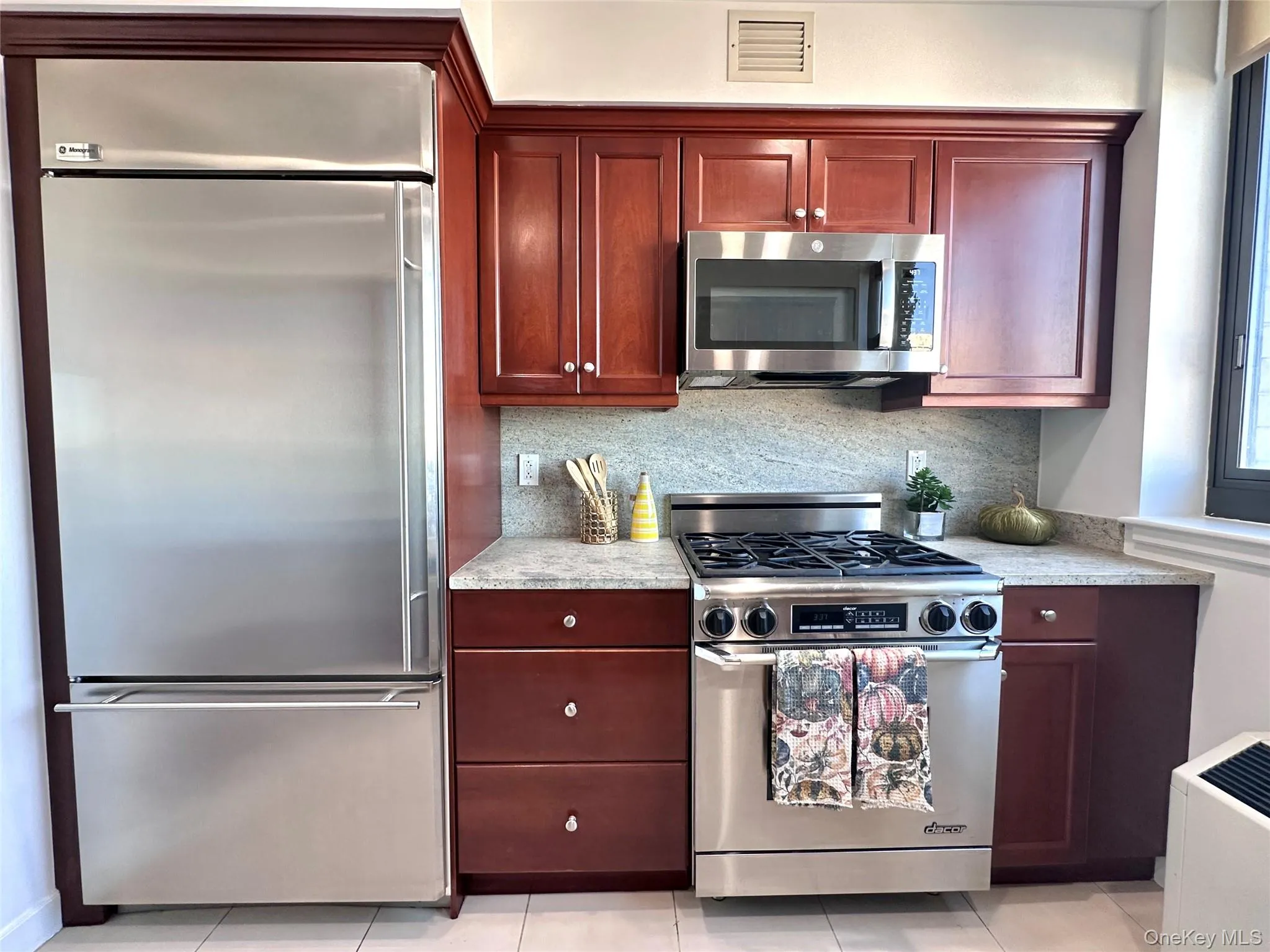 Kitchen featuring high quality appliances, tasteful backsplash, light tile patterned floors, granite counters, and reddish brown cabinets Kitchen featuring high quality appliances, tasteful backsplash, light tile patterned floors, granite counters, and reddish brown cabinets