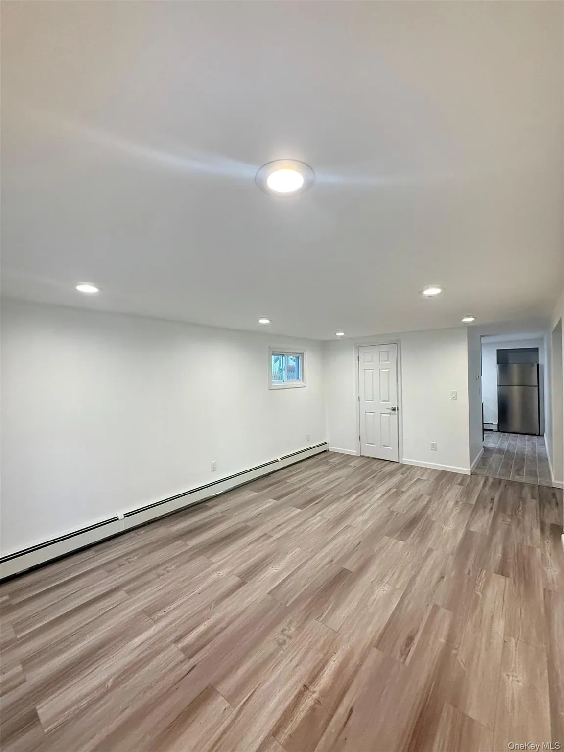 Basement featuring a baseboard radiator, light wood-style flooring, freestanding refrigerator, and recessed lighting Basement featuring a baseboard radiator, light wood-style flooring, freestanding refrigerator, and recessed lighting