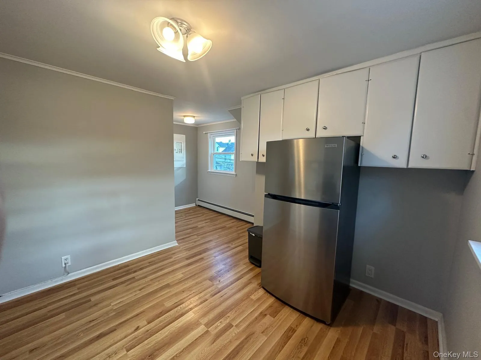 Kitchen featuring freestanding refrigerator, white cabinetry, light wood-style flooring, a baseboard heating unit, and ornamental molding Kitchen featuring freestanding refrigerator, white cabinetry, light wood-style flooring, a baseboard heating unit, and ornamental molding