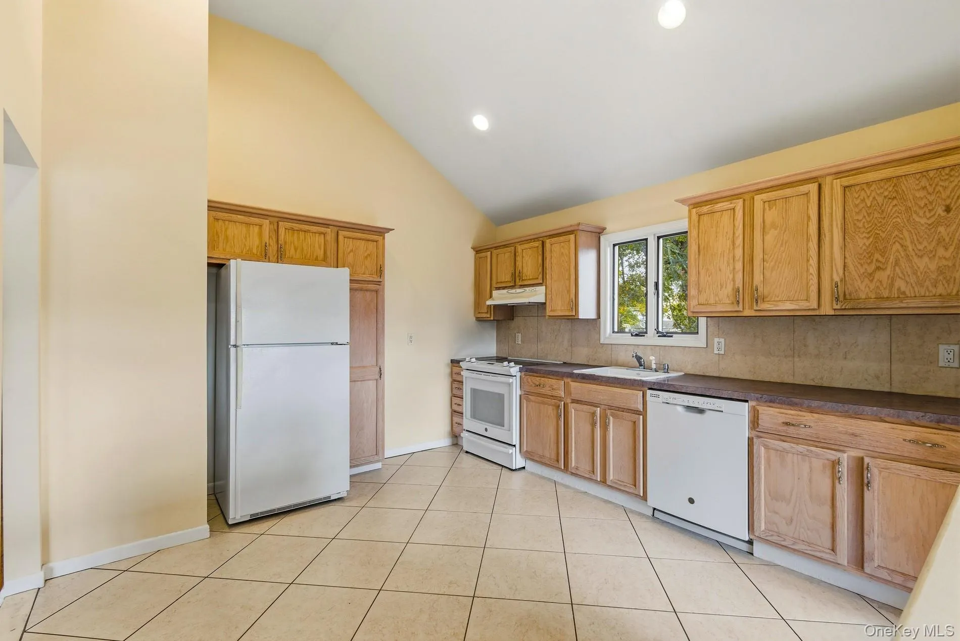 Kitchen featuring white appliances, dark countertops, recessed lighting, light tile patterned flooring, and backsplash Kitchen featuring white appliances, dark countertops, recessed lighting, light tile patterned flooring, and backsplash