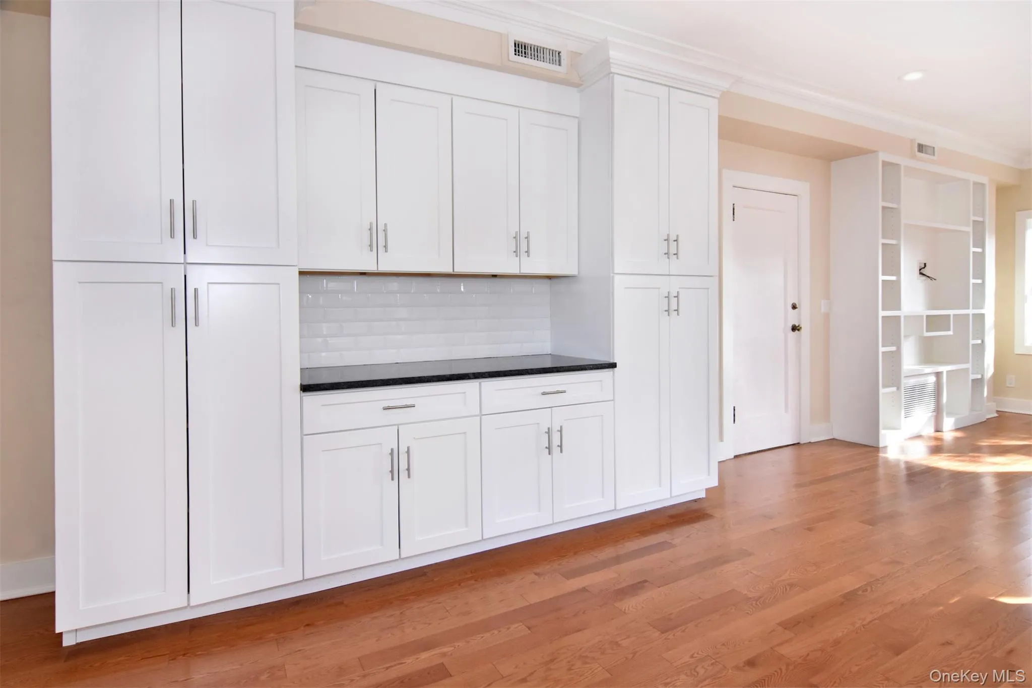 Kitchen with white cabinets, light wood finished floors, crown molding, tasteful backsplash, and dark stone counters Kitchen with white cabinets, light wood finished floors, crown molding, tasteful backsplash, and dark stone counters