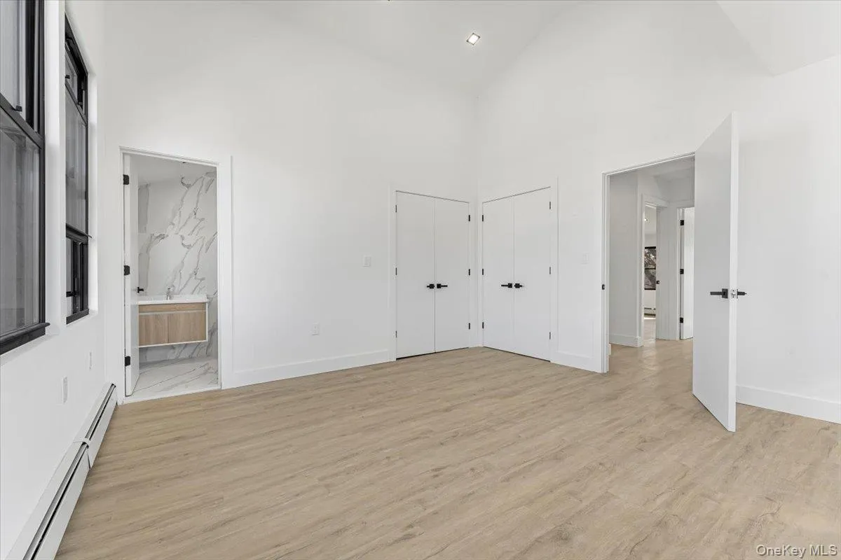 Unfurnished bedroom with high vaulted ceiling, baseboard heating, multiple closets, light wood-style floors, and ensuite bathroom Unfurnished bedroom with high vaulted ceiling, baseboard heating, multiple closets, light wood-style floors, and ensuite bathroom