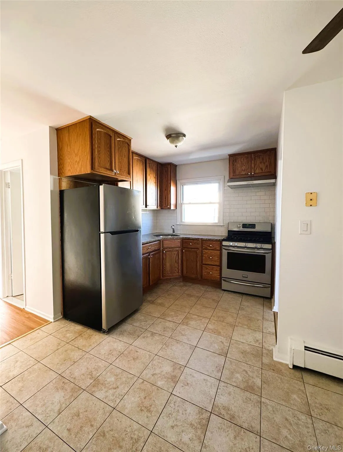 Kitchen with appliances with stainless steel finishes, a baseboard heating unit, light tile patterned floors, brown cabinetry, and backsplash Kitchen with appliances with stainless steel finishes, a baseboard heating unit, light tile patterned floors, brown cabinetry, and backsplash