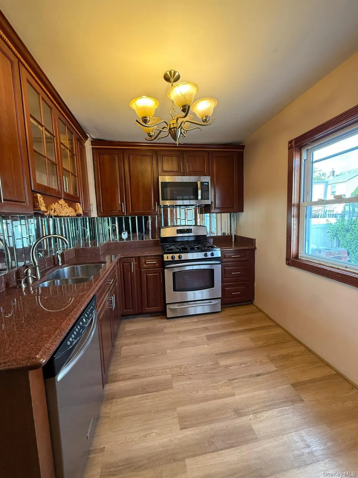 Kitchen featuring appliances with stainless steel finishes, glass insert cabinets, a chandelier, dark stone counters, and light wood finished floors Kitchen featuring appliances with stainless steel finishes, glass insert cabinets, a chandelier, dark stone counters, and light wood finished floors