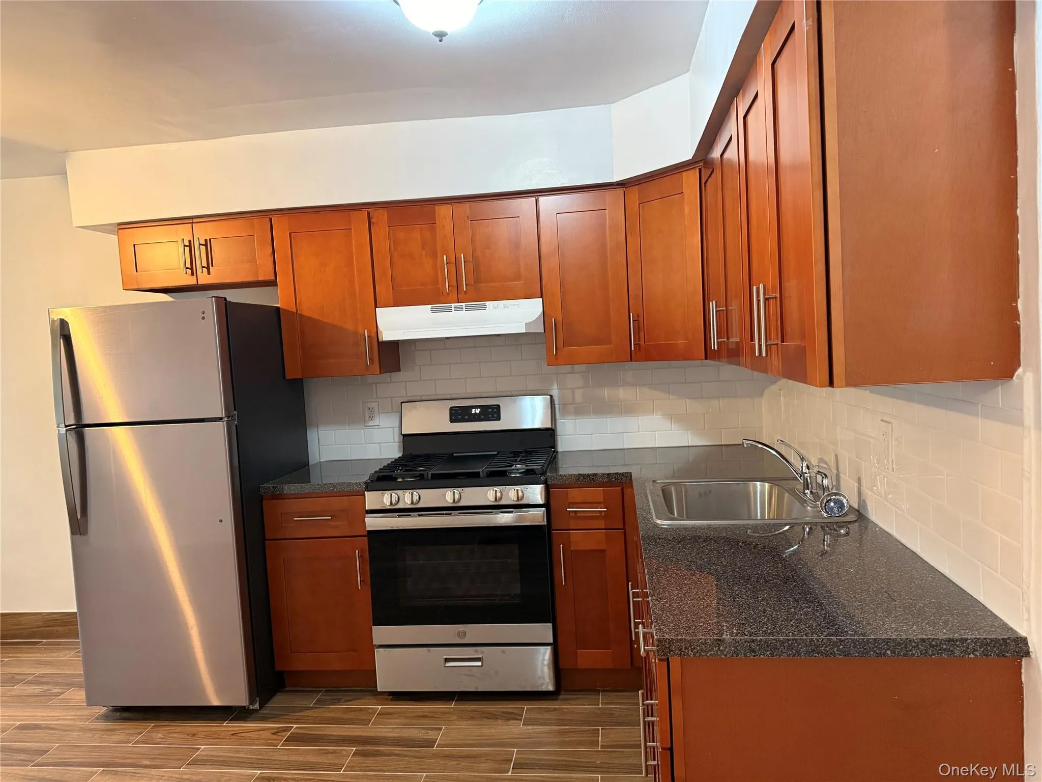 Kitchen featuring stainless steel appliances, backsplash, wood finish floors, dark stone counters, and brown cabinets Kitchen featuring stainless steel appliances, backsplash, wood finish floors, dark stone counters, and brown cabinets