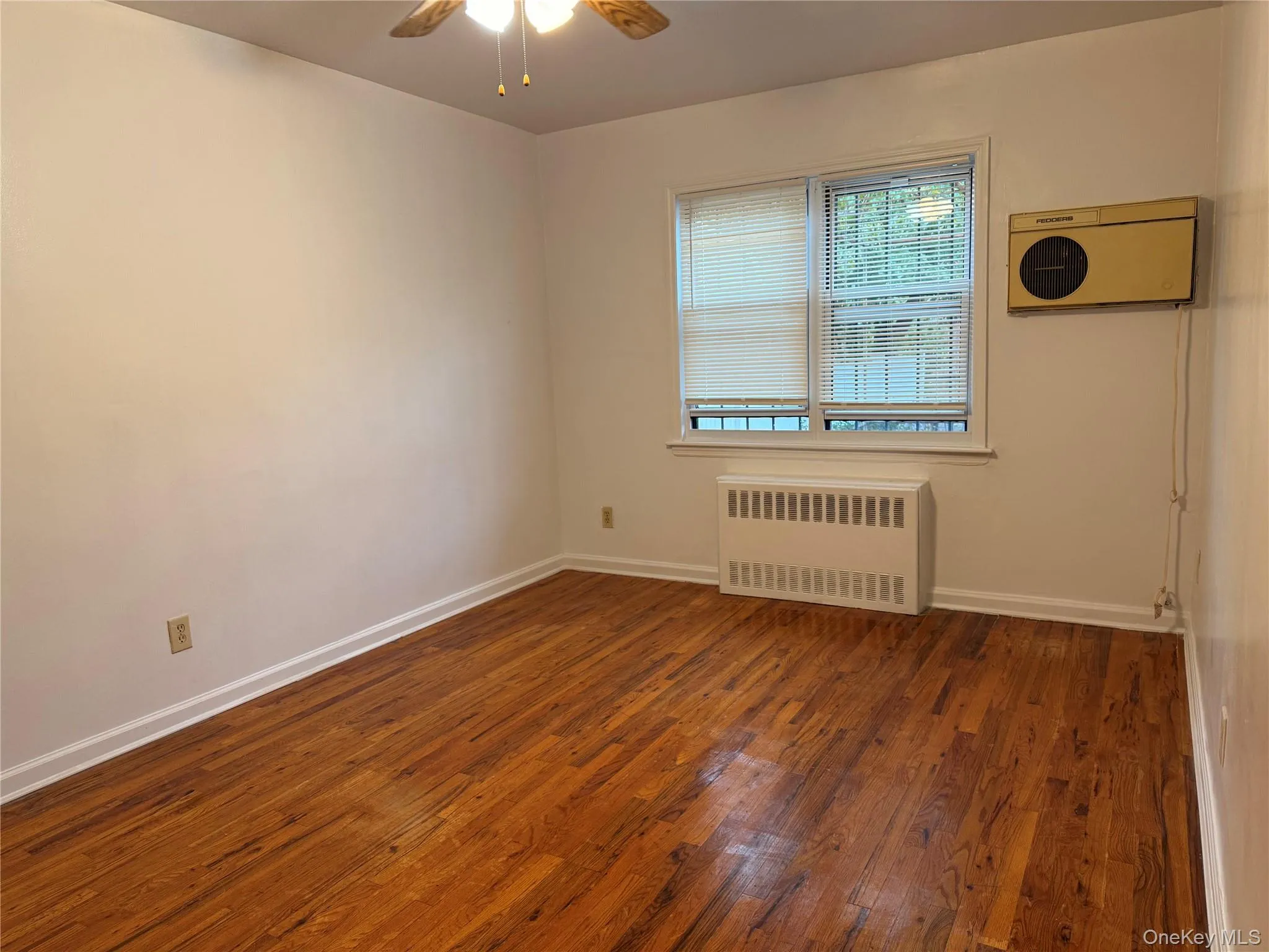 Empty room featuring dark wood-type flooring, radiator heating unit, and a ceiling fan Empty room featuring dark wood-type flooring, radiator heating unit, and a ceiling fan
