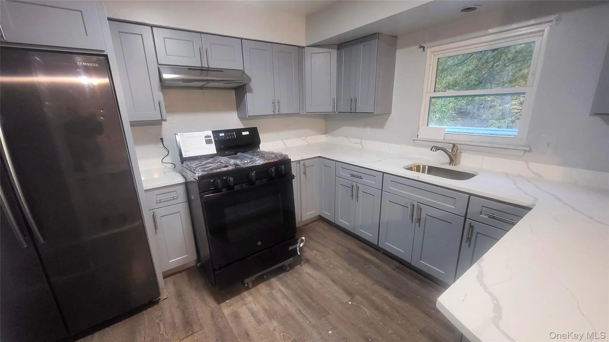 Kitchen with gray cabinetry, freestanding refrigerator, black range with gas cooktop, light stone countertops, and dark wood finished floors Kitchen with gray cabinetry, freestanding refrigerator, black range with gas cooktop, light stone countertops, and dark wood finished floors