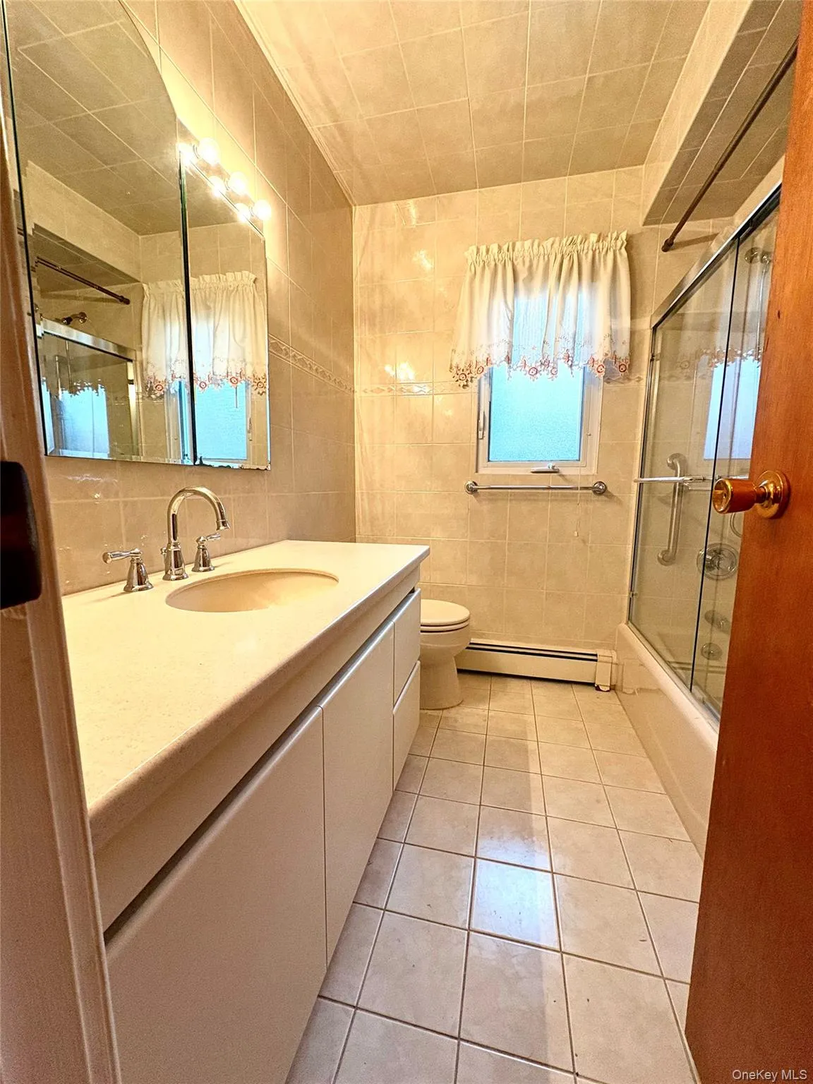 Full bath featuring tile walls, light tile patterned floors, vanity, shower / bath combination with glass door, and a baseboard radiator Full bath featuring tile walls, light tile patterned floors, vanity, shower / bath combination with glass door, and a baseboard radiator