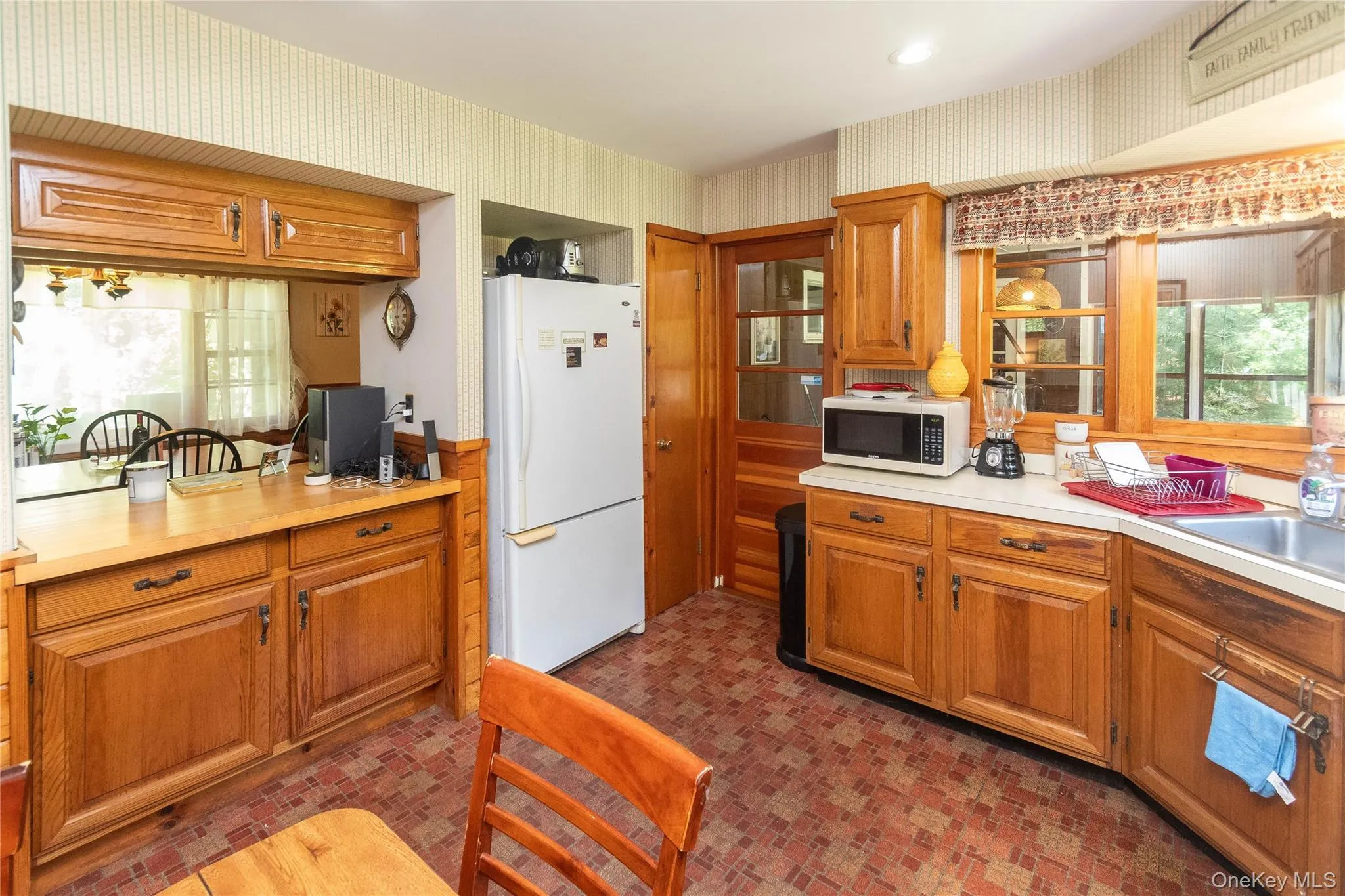 Kitchen with freestanding refrigerator, wallpapered walls, light countertops, stainless steel microwave, and brown cabinetry Kitchen with freestanding refrigerator, wallpapered walls, light countertops, stainless steel microwave, and brown cabinetry