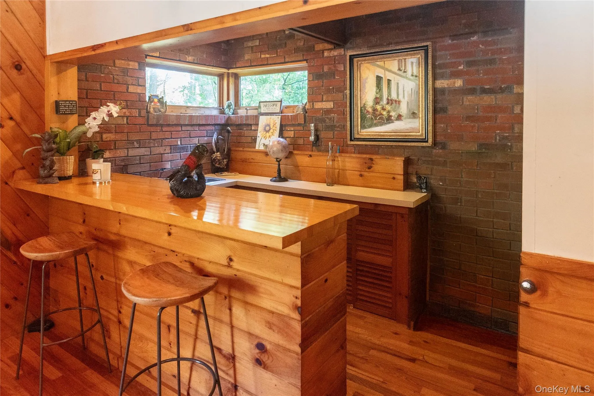 Indoor dry bar featuring light wood-style floors, brick wall, wood counters, and brown cabinetry Indoor dry bar featuring light wood-style floors, brick wall, wood counters, and brown cabinetry