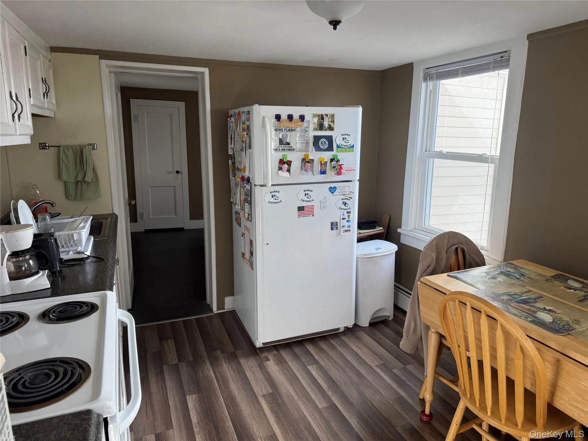 Kitchen with white appliances, dark wood finished floors, white cabinets, dark countertops, and a baseboard radiator Kitchen with white appliances, dark wood finished floors, white cabinets, dark countertops, and a baseboard radiator