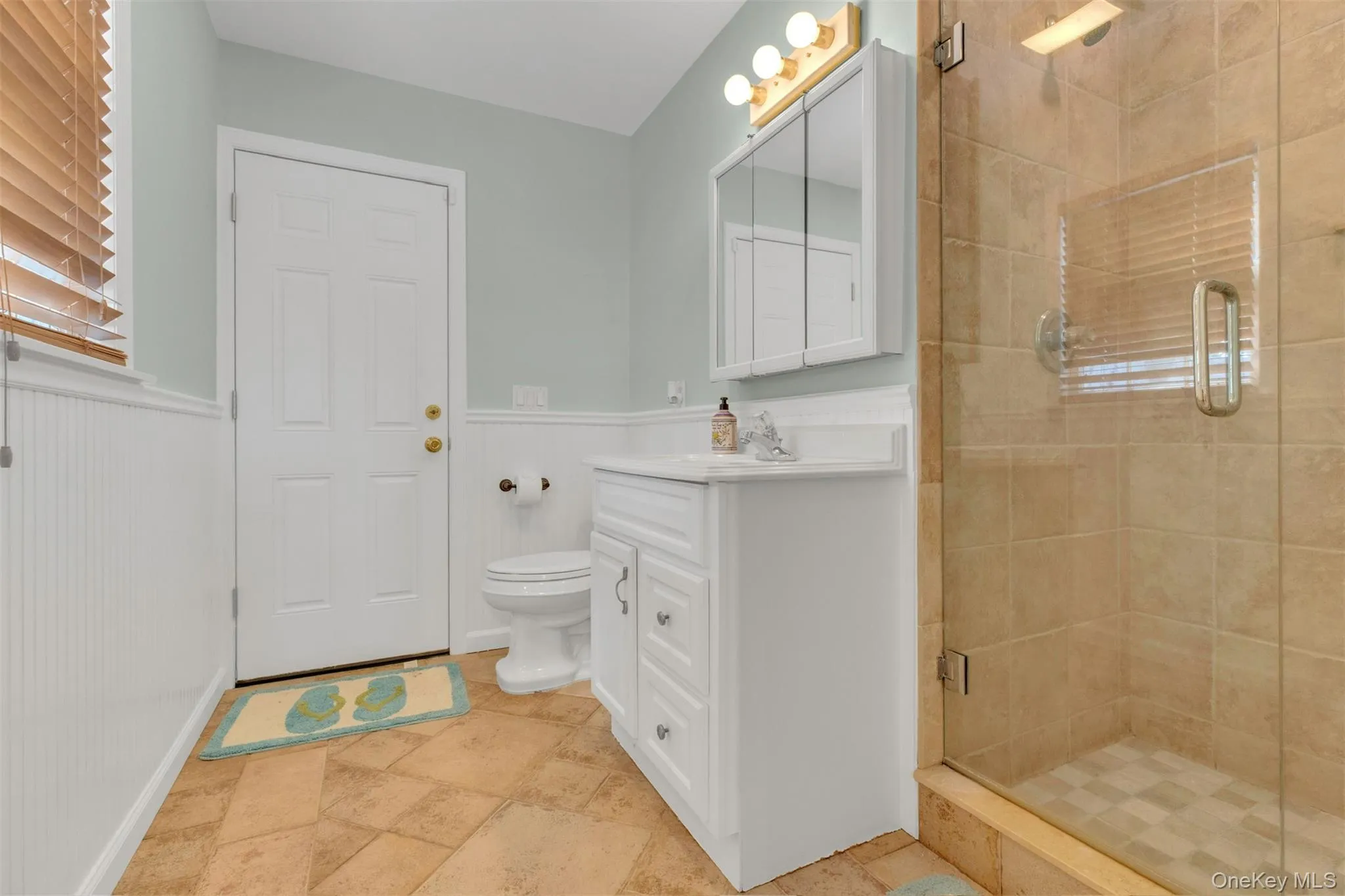 Bathroom with a shower stall, vanity, stone tile flooring, and a wainscoted wall Bathroom with a shower stall, vanity, stone tile flooring, and a wainscoted wall