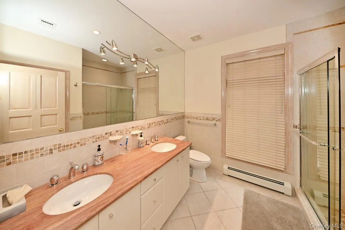 Bathroom featuring tile walls, a baseboard radiator, light tile patterned flooring, a stall shower, and double vanity Bathroom featuring tile walls, a baseboard radiator, light tile patterned flooring, a stall shower, and double vanity