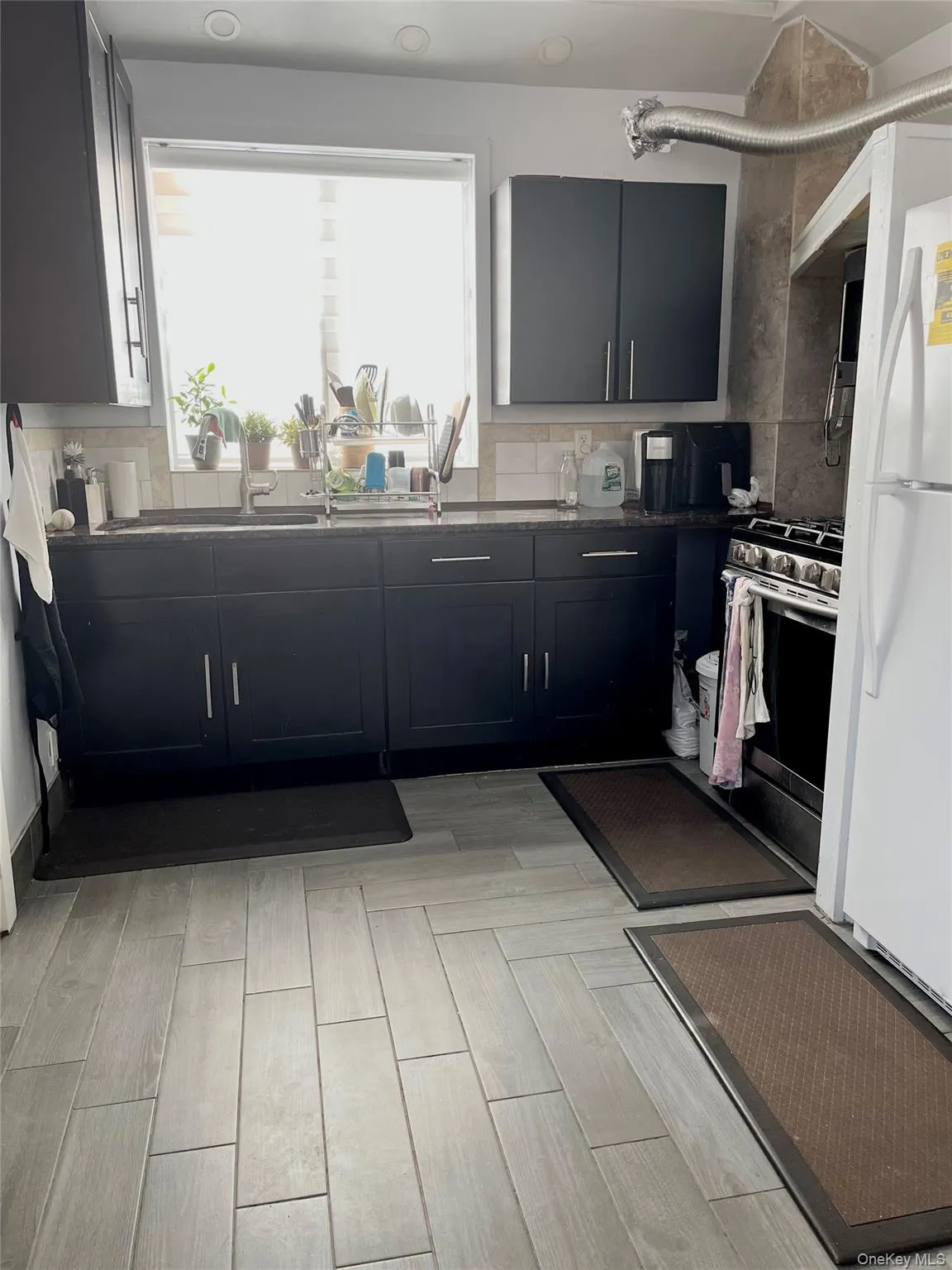 Kitchen featuring dark cabinetry, decorative backsplash, freestanding refrigerator, stainless steel gas range, and light wood-style flooring Kitchen featuring dark cabinetry, decorative backsplash, freestanding refrigerator, stainless steel gas range, and light wood-style flooring