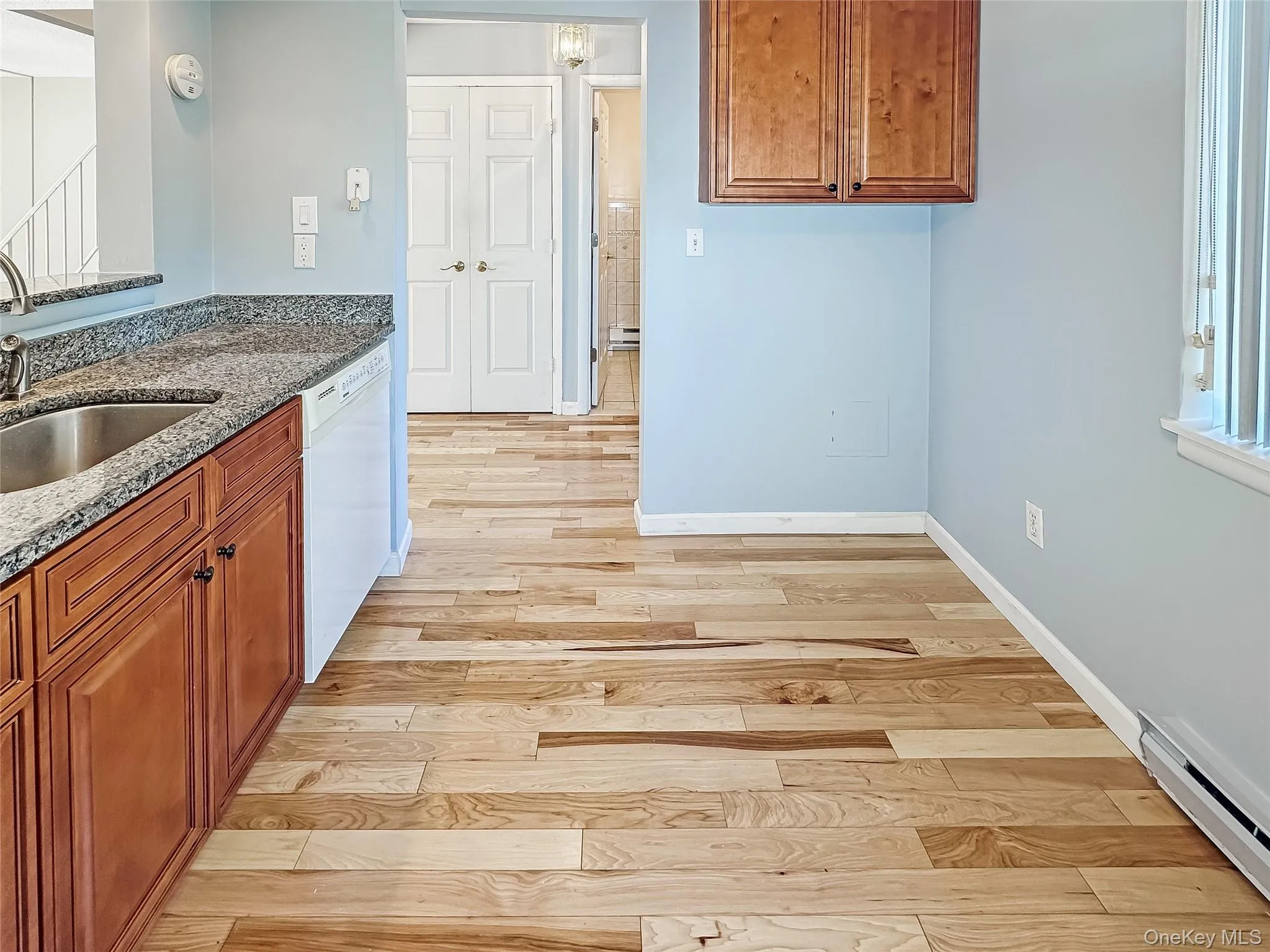 Kitchen featuring dark stone countertops, brown cabinetry, light wood-type flooring, a baseboard radiator, and dishwasher Kitchen featuring dark stone countertops, brown cabinetry, light wood-type flooring, a baseboard radiator, and dishwasher
