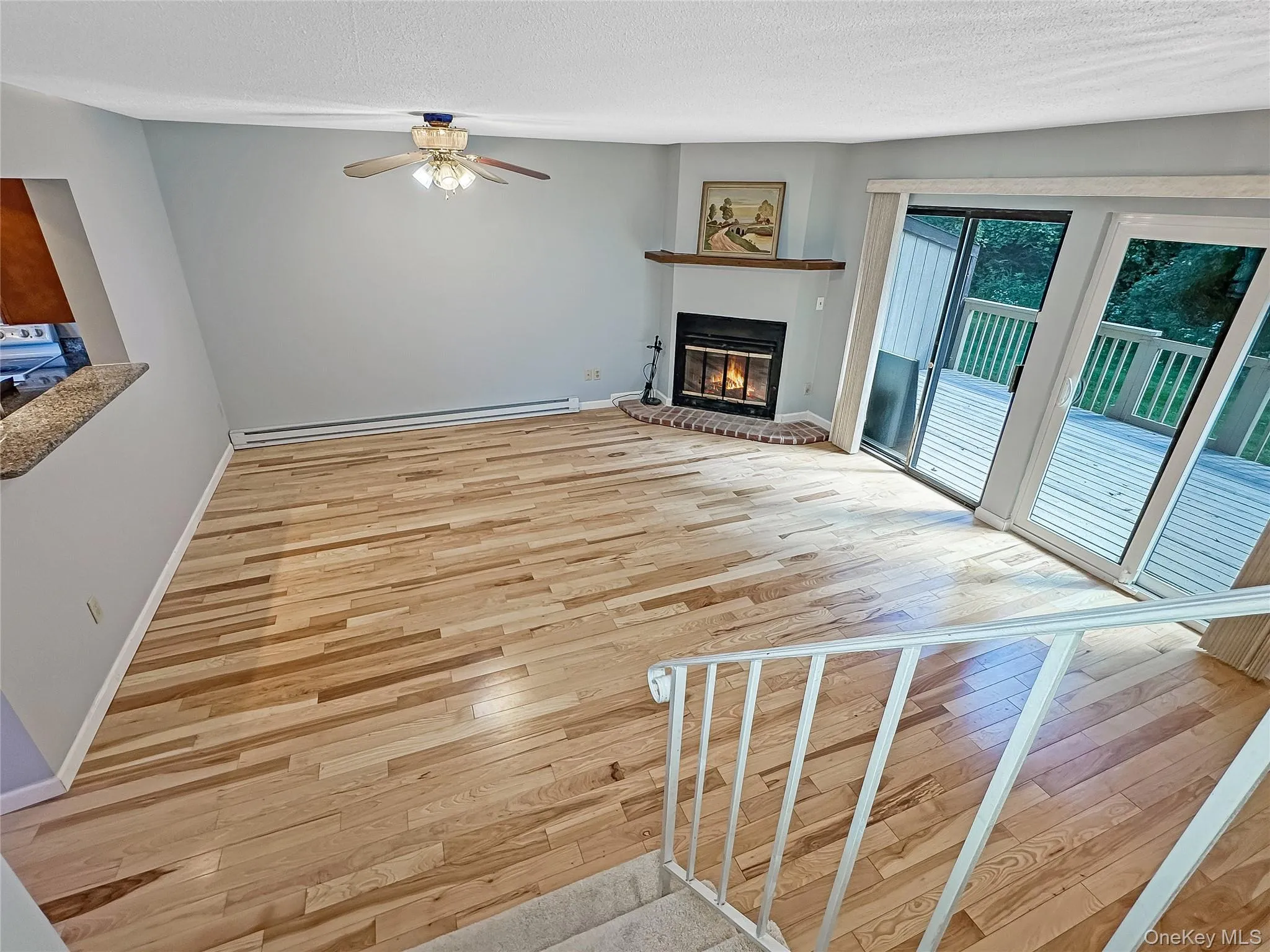 Unfurnished living room with light wood-style flooring, a glass covered fireplace, a baseboard heating unit, a textured ceiling, and ceiling fan Unfurnished living room with light wood-style flooring, a glass covered fireplace, a baseboard heating unit, a textured ceiling, and ceiling fan