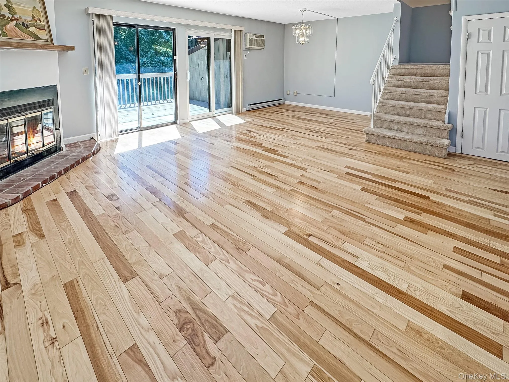 Unfurnished living room with light wood finished floors, stairs, a brick fireplace, a chandelier, and baseboard heating Unfurnished living room with light wood finished floors, stairs, a brick fireplace, a chandelier, and baseboard heating