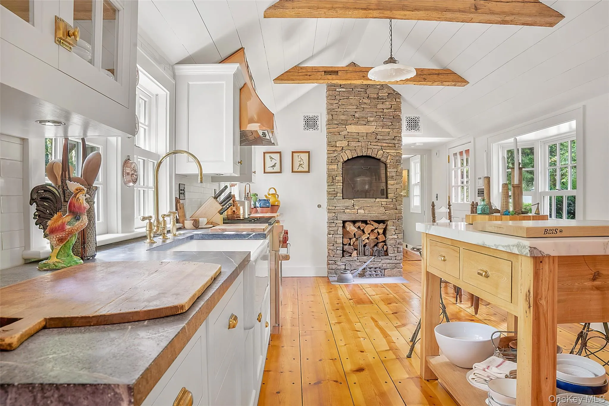 Kitchen with white cabinets, plenty of natural light, light wood-style flooring, a stone fireplace, and beamed ceiling Kitchen with white cabinets, plenty of natural light, light wood-style flooring, a stone fireplace, and beamed ceiling