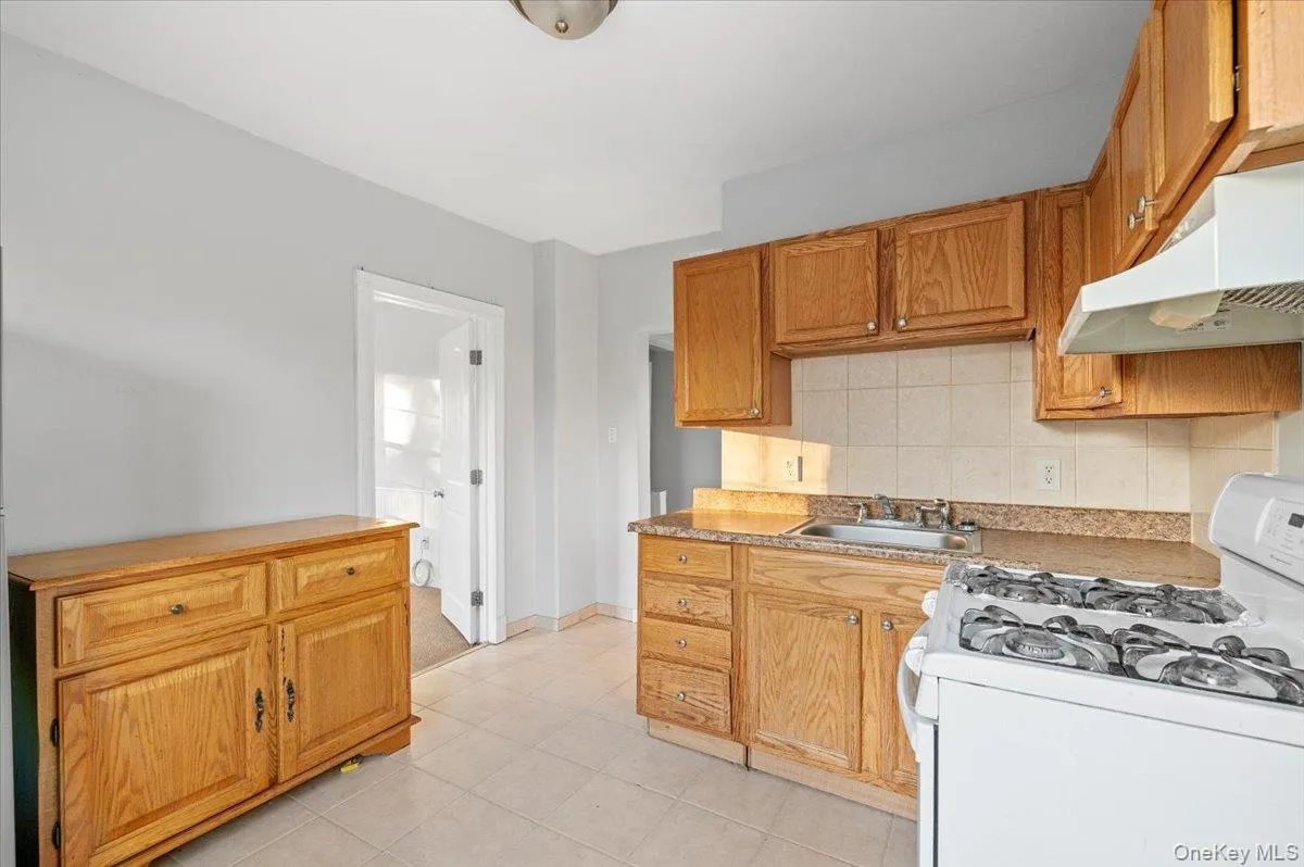 Kitchen featuring white gas range, backsplash, under cabinet range hood, brown cabinetry, and light countertops Kitchen featuring white gas range, backsplash, under cabinet range hood, brown cabinetry, and light countertops